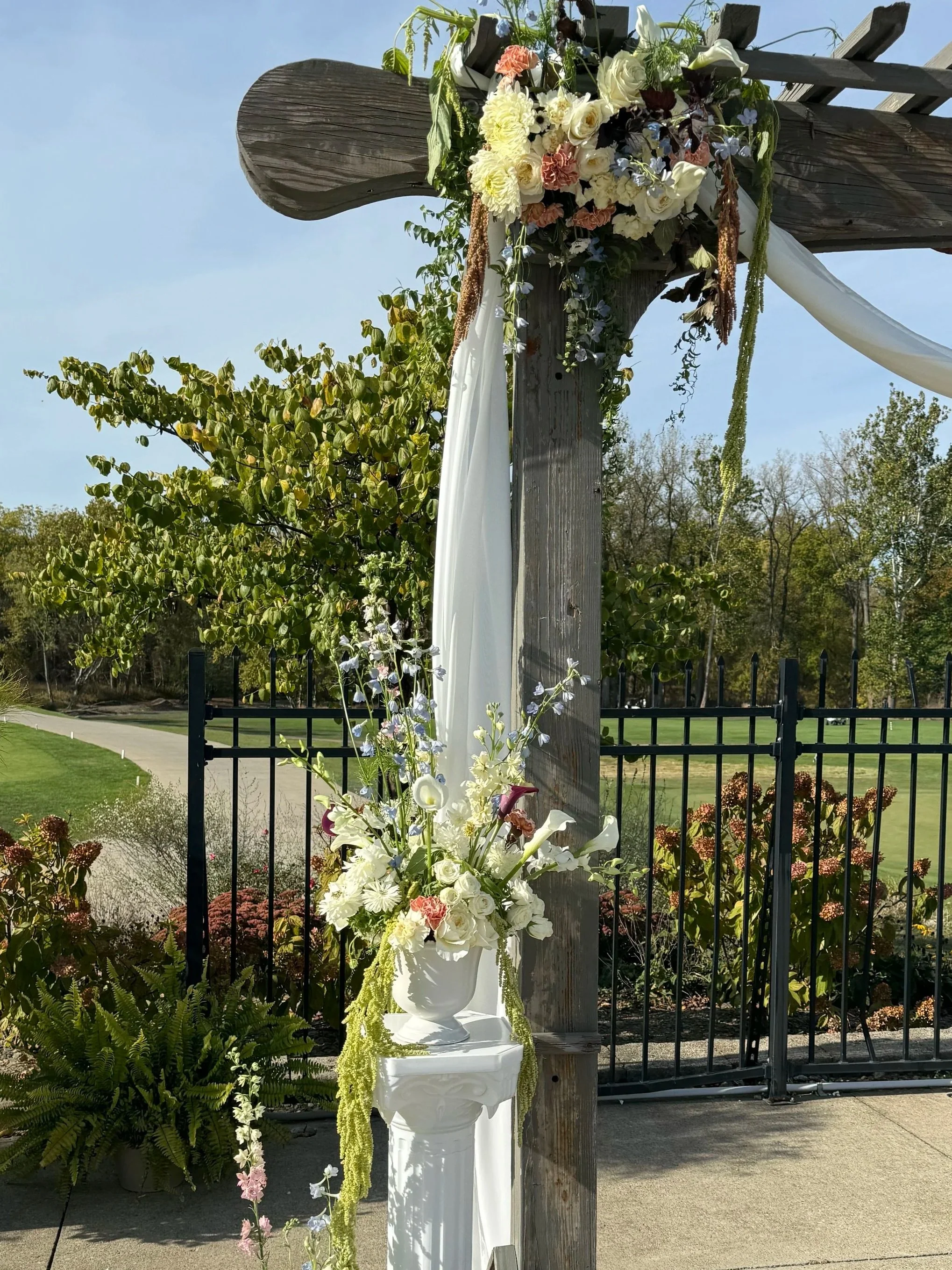 A wedding arch decorated with hanging flowers, white drapery, and a floral arrangement on a white pedestal outdoors.