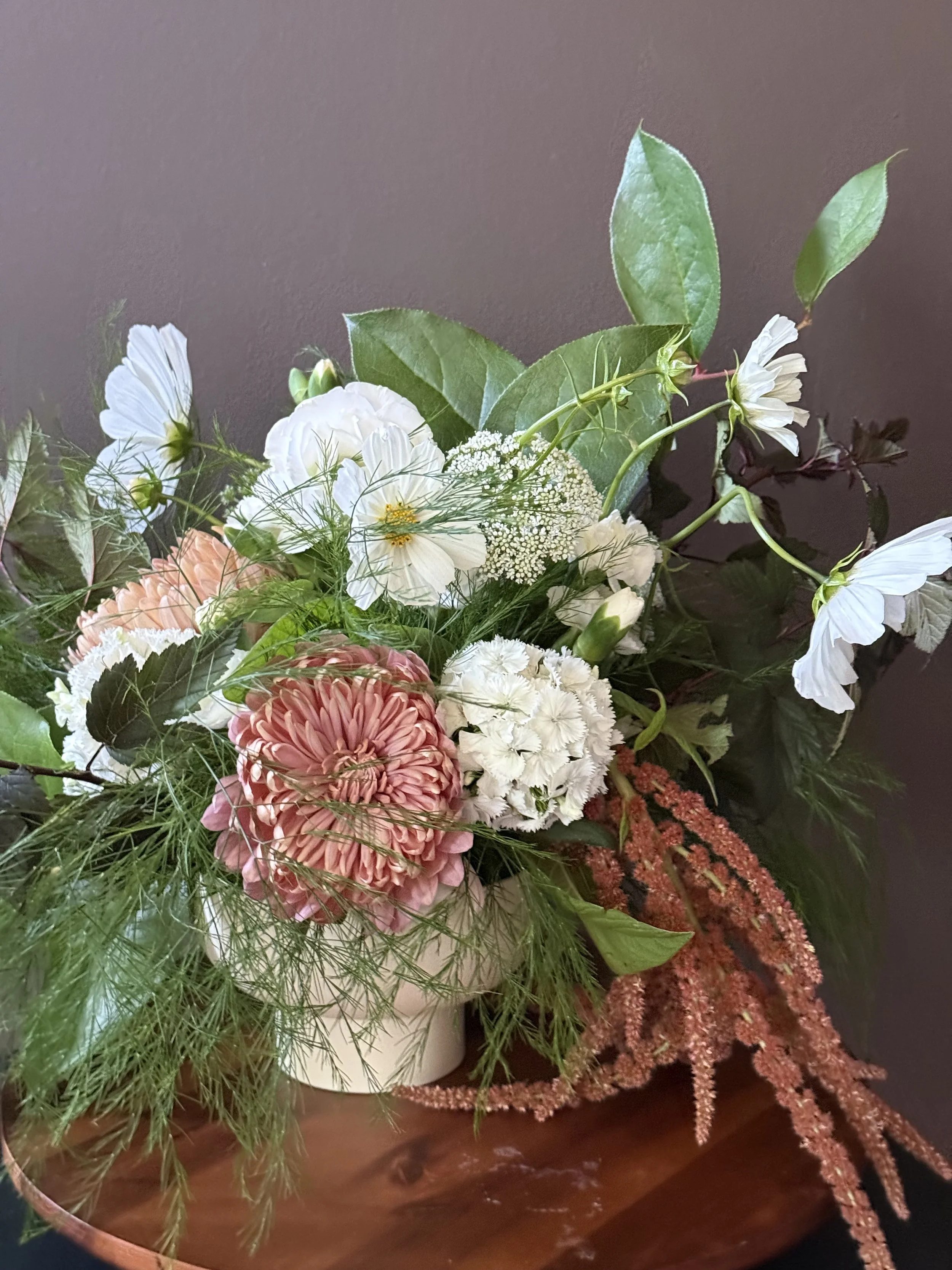 A floral arrangement with white, pink, and green flowers and greenery in a white vase, set on a wooden surface against a dark wall.