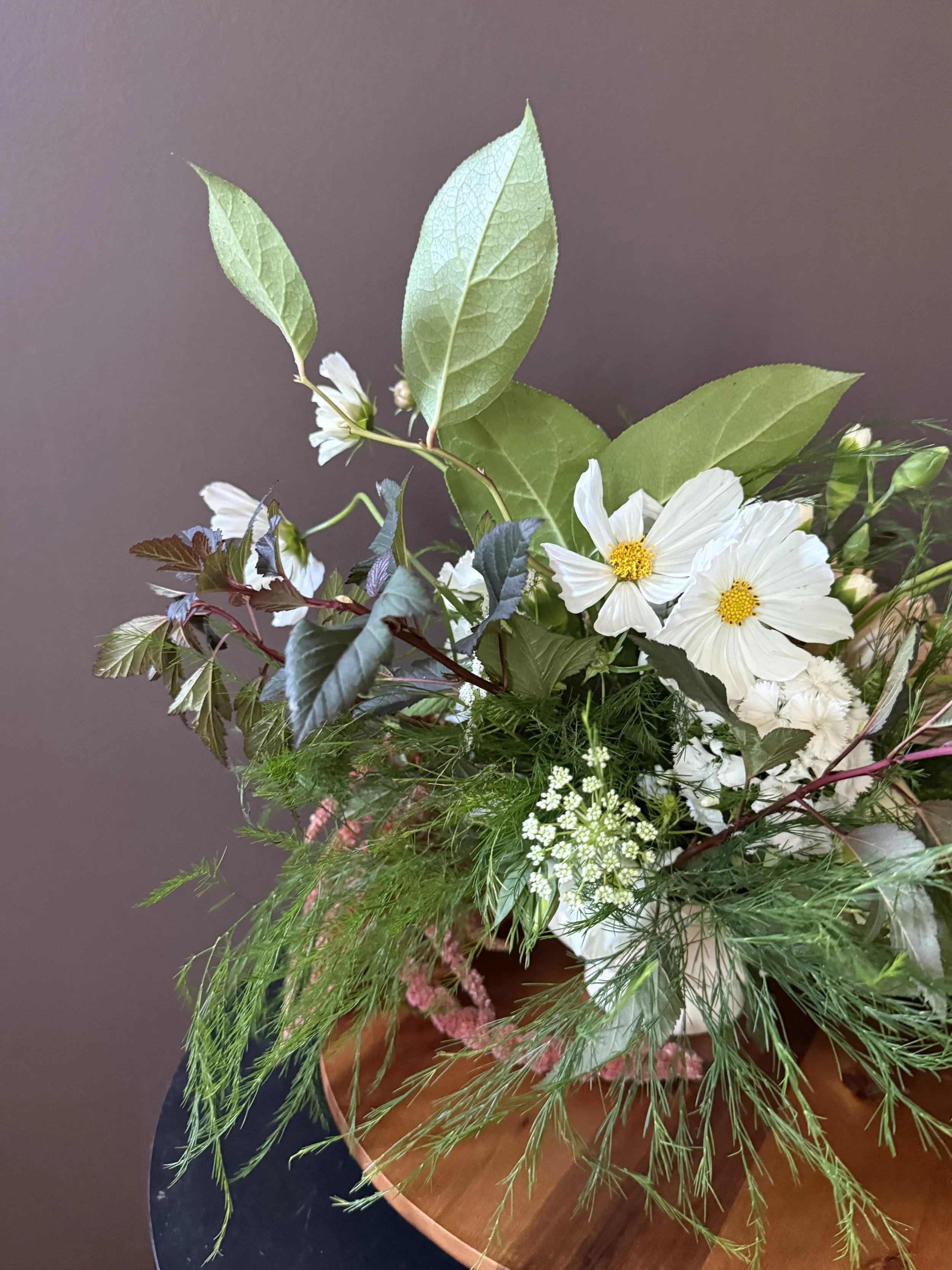 A bouquet of white flowers and green leaves in a wooden bowl on a dark surface.