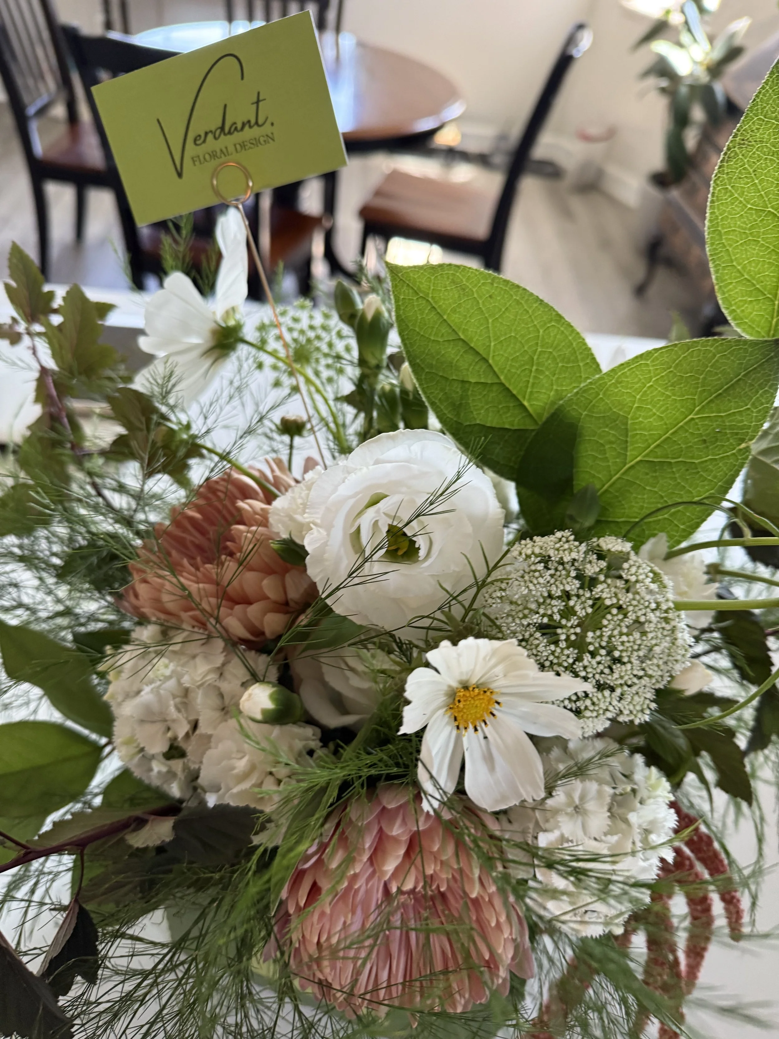 Close-up of a floral arrangement with white, pink, and green flowers, including daisies and other blossoms, with a yellow business card labeled "Verdant Floral Design" in the background.