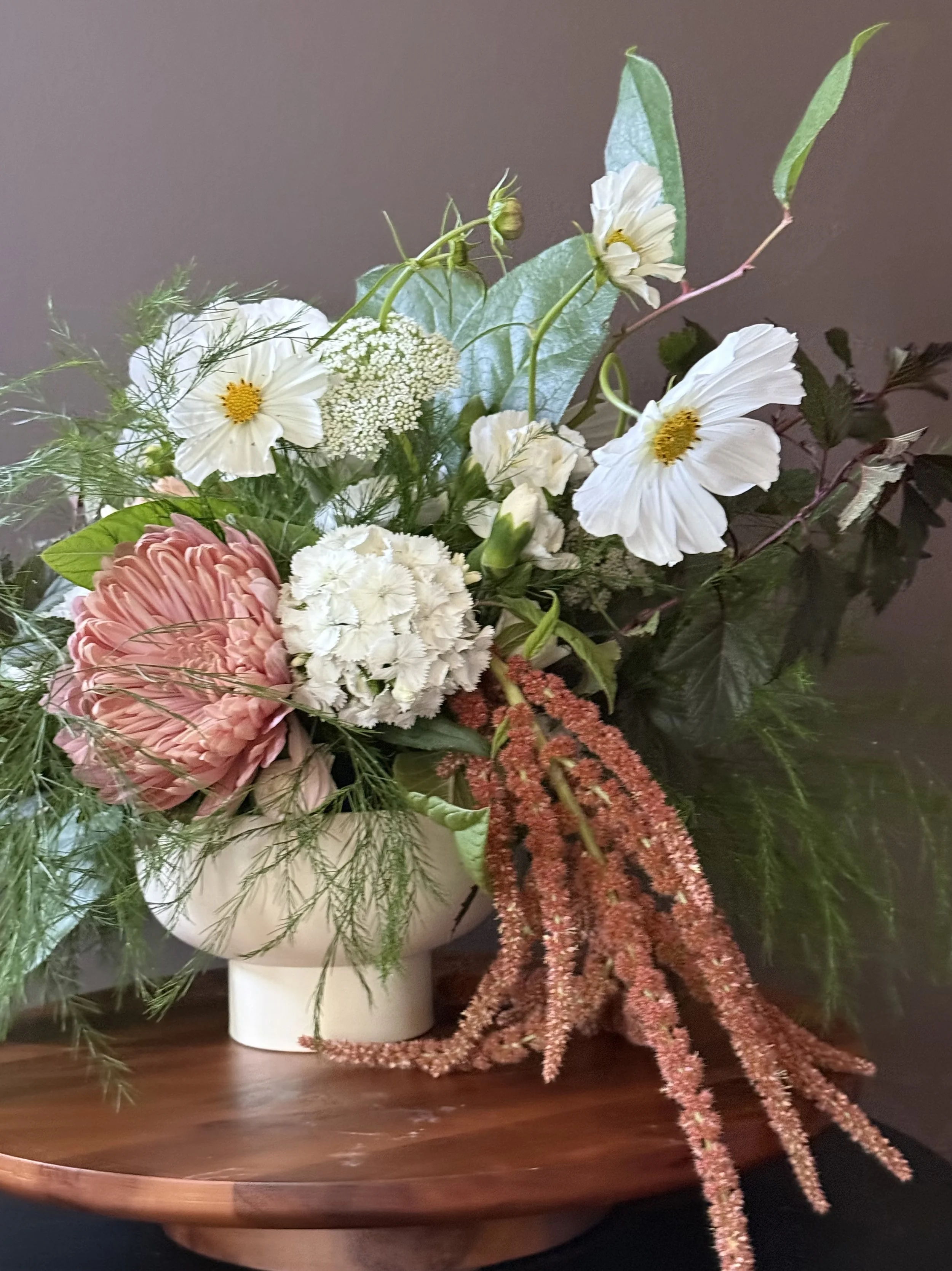 A floral arrangement in a white vase containing pink, white, and yellow flowers, green leaves, and long red-brown stalks, placed on a wooden surface.