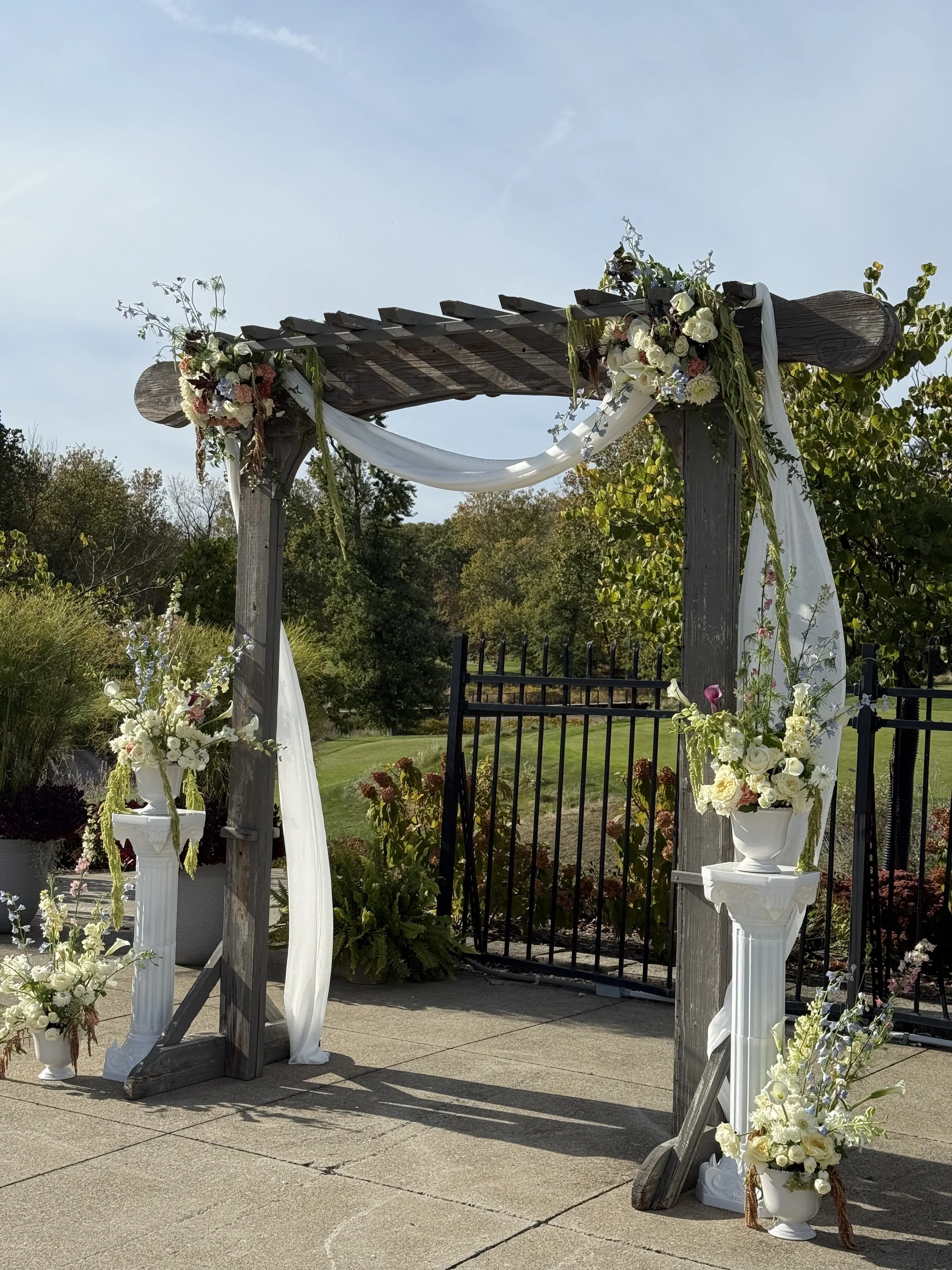 Wooden wedding arch decorated with white and pink flowers, white drapes, and greenery, set outdoors with a garden background.