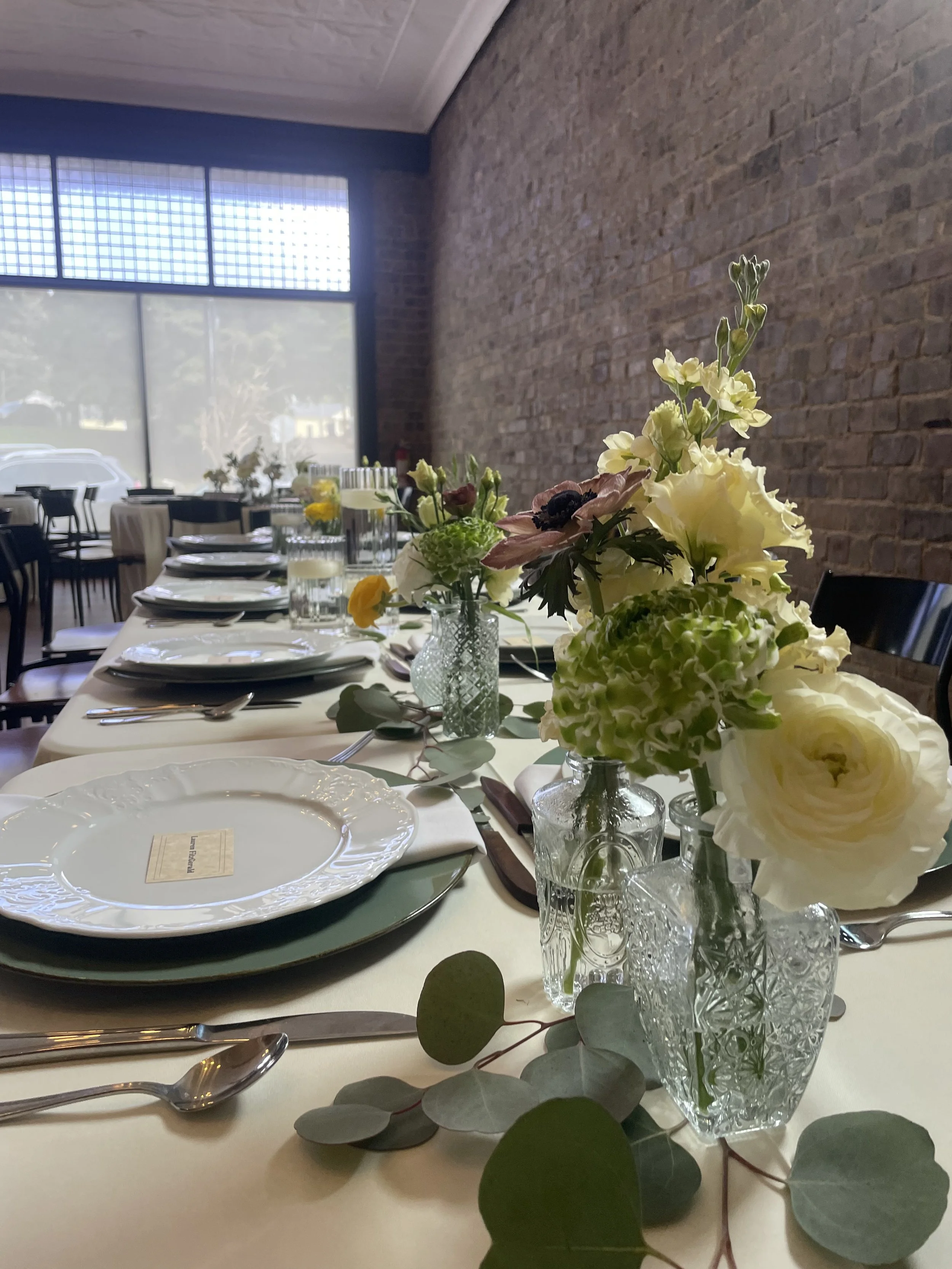 A long dining table decorated with floral centerpieces, plates, and cutlery in a restaurant with brick walls and large windows.