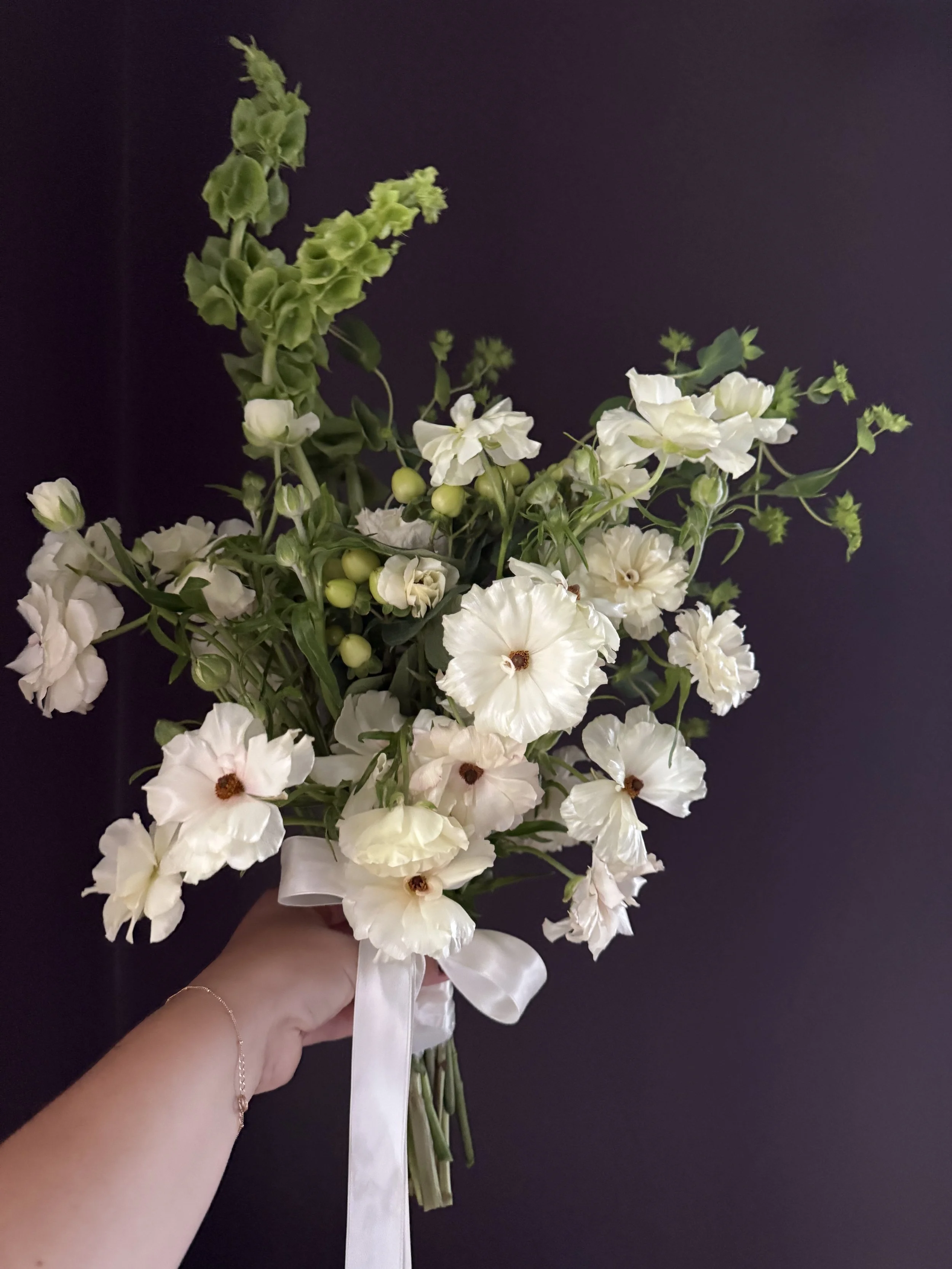 A hand holding a bouquet of white flowers with green stems and leaves, tied with a white ribbon, against a dark background.