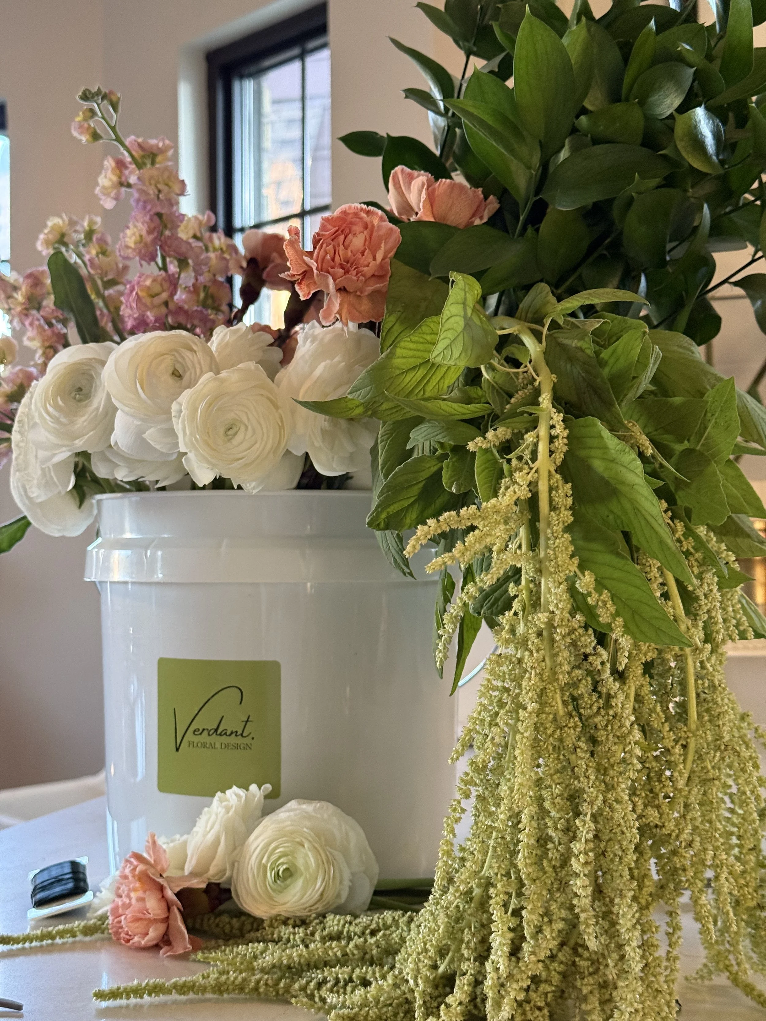Arrangement of various flowers, including white ranunculus, pink carnations, and greenery, inside a white container labeled 'Verdant Floral Design' with some flowers and stems lying on the surface.