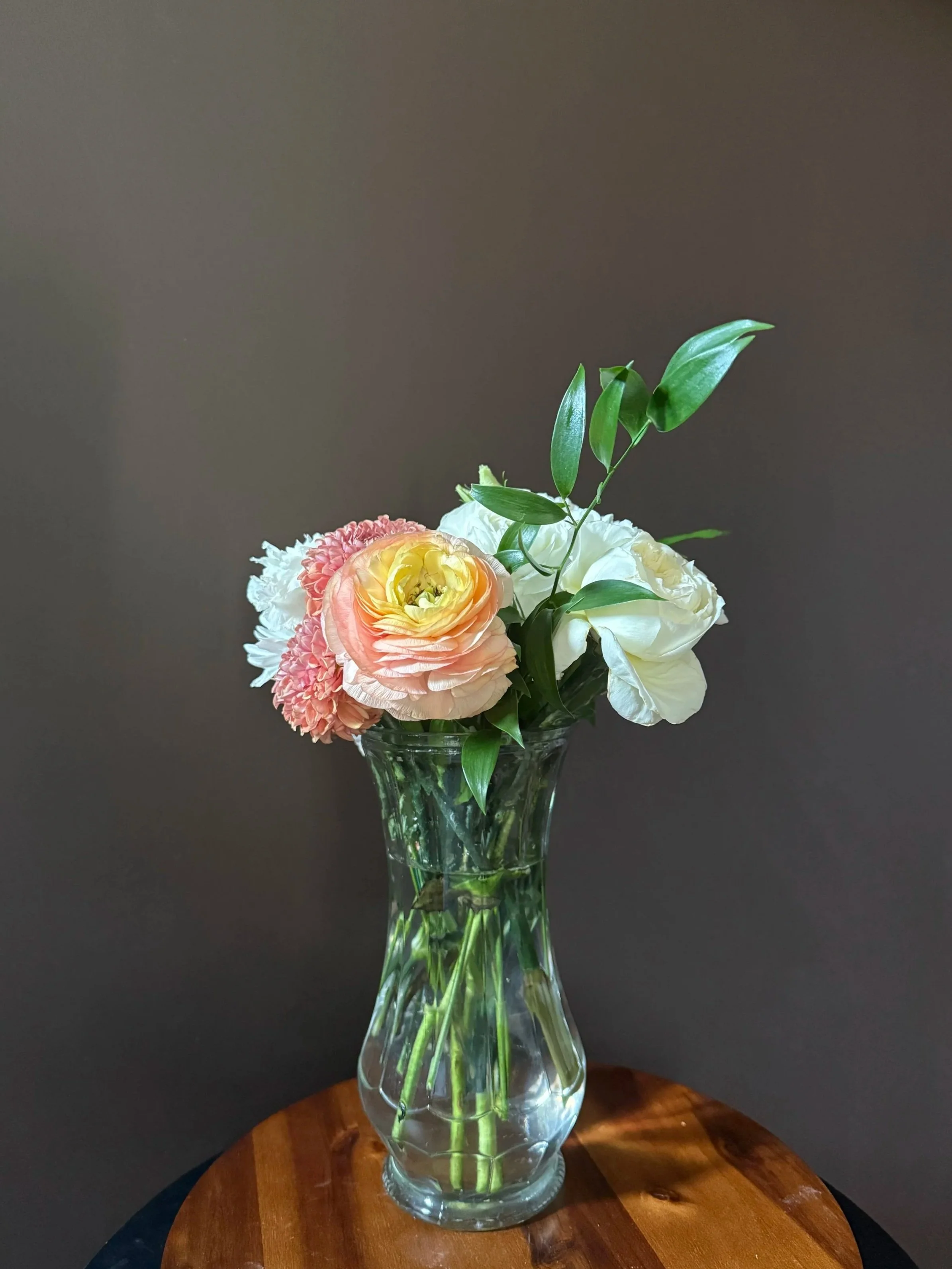 A glass vase with water holding a bouquet of pink, white, and yellow flowers and green foliage on a wooden surface against a gray background.