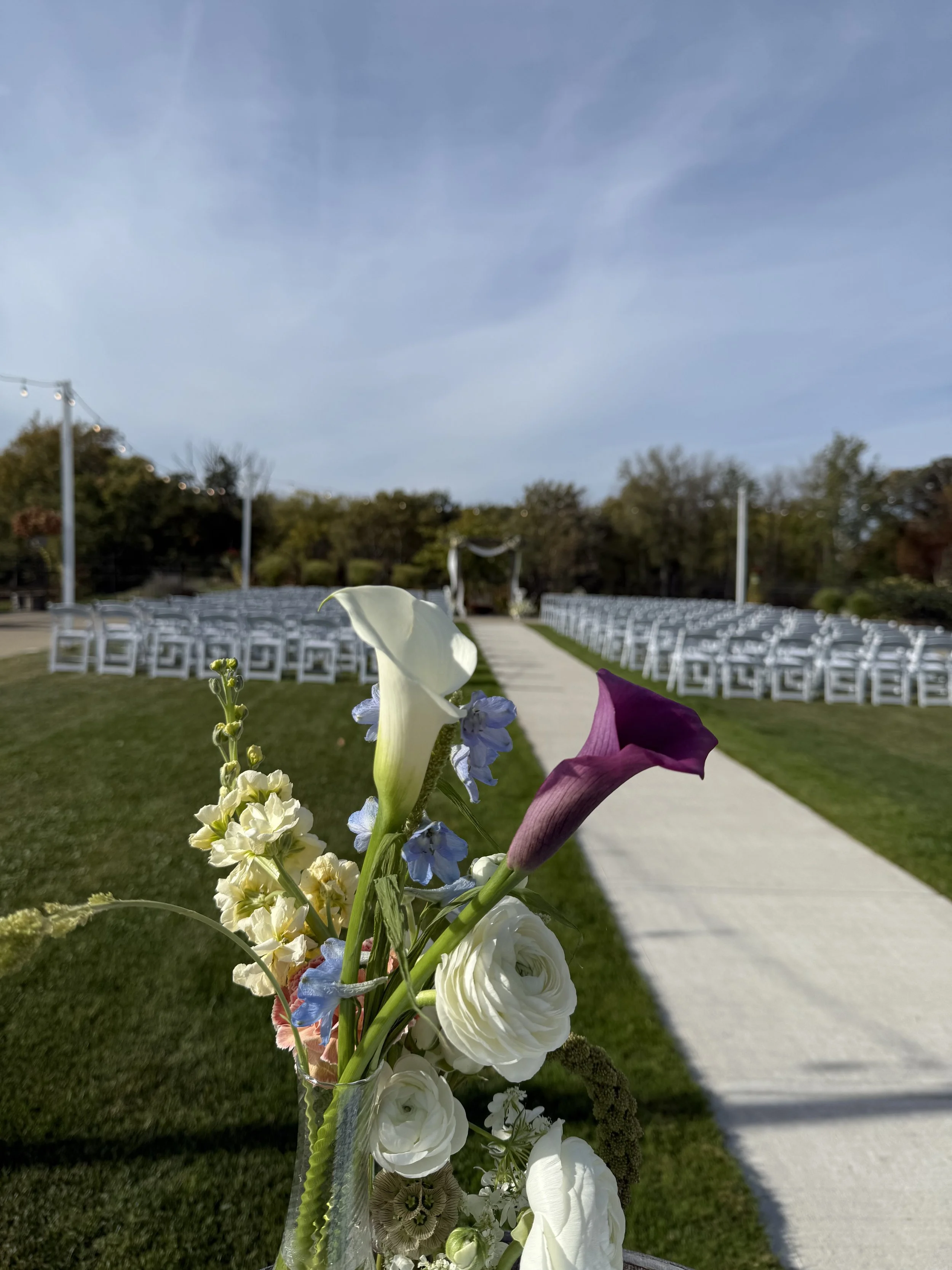 A wedding aisle with white chairs on both sides and a floral arrangement in the foreground. The aisle leads to a wedding arch under a partly cloudy sky.
