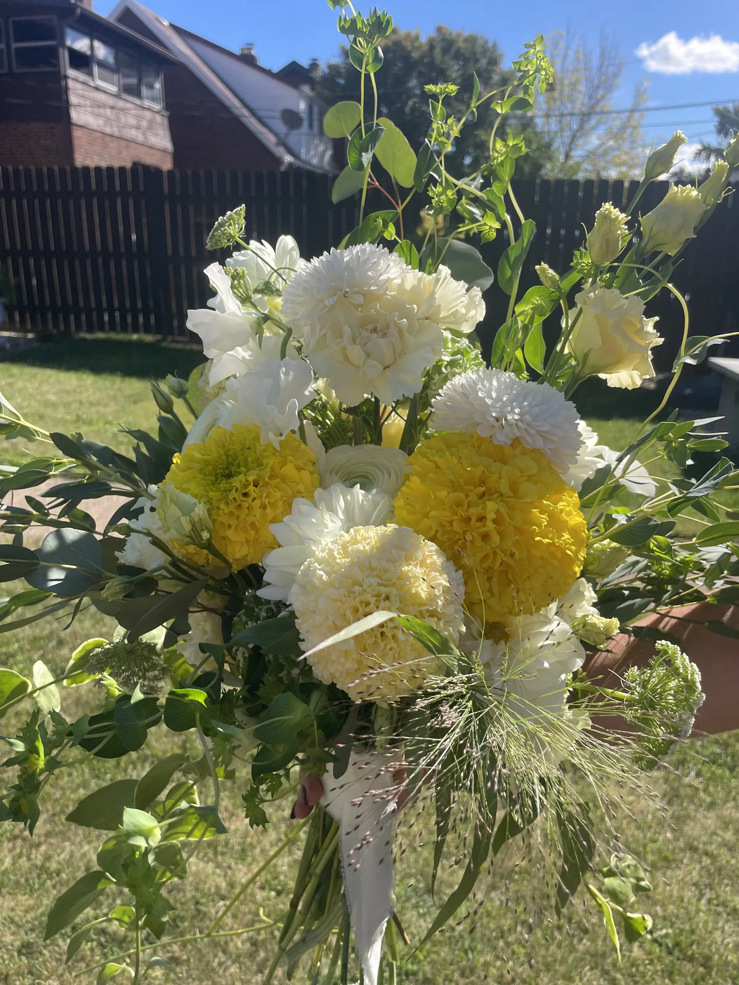 A bouquet of white and yellow flowers held outdoors on a sunny day with a grassy yard, a black fence, and houses in the background.