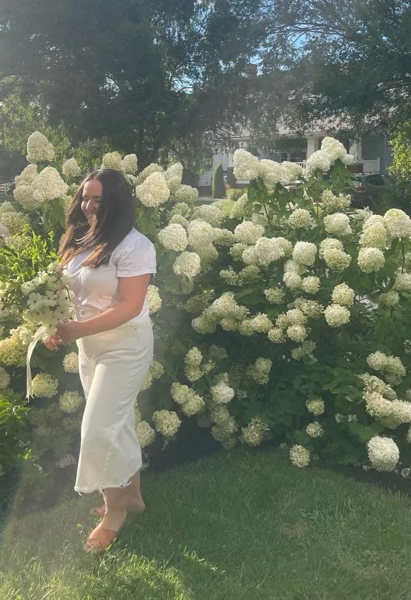 A woman in white standing barefoot on grass, holding a bouquet of white flowers, in front of white hydrangea bushes in a garden.