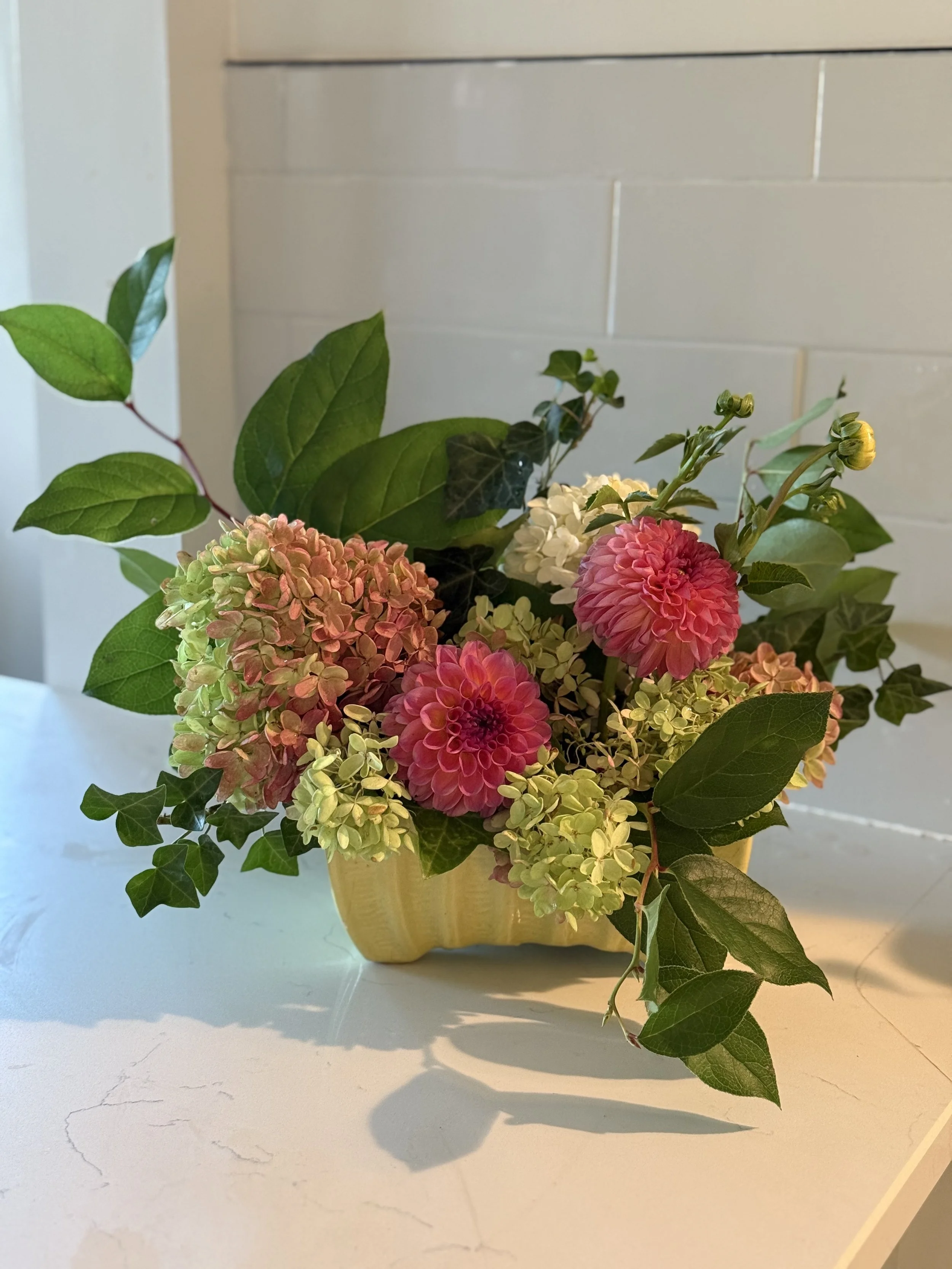 A floral arrangement with pink and white flowers and green foliage in a yellow rectangular container on a white surface.