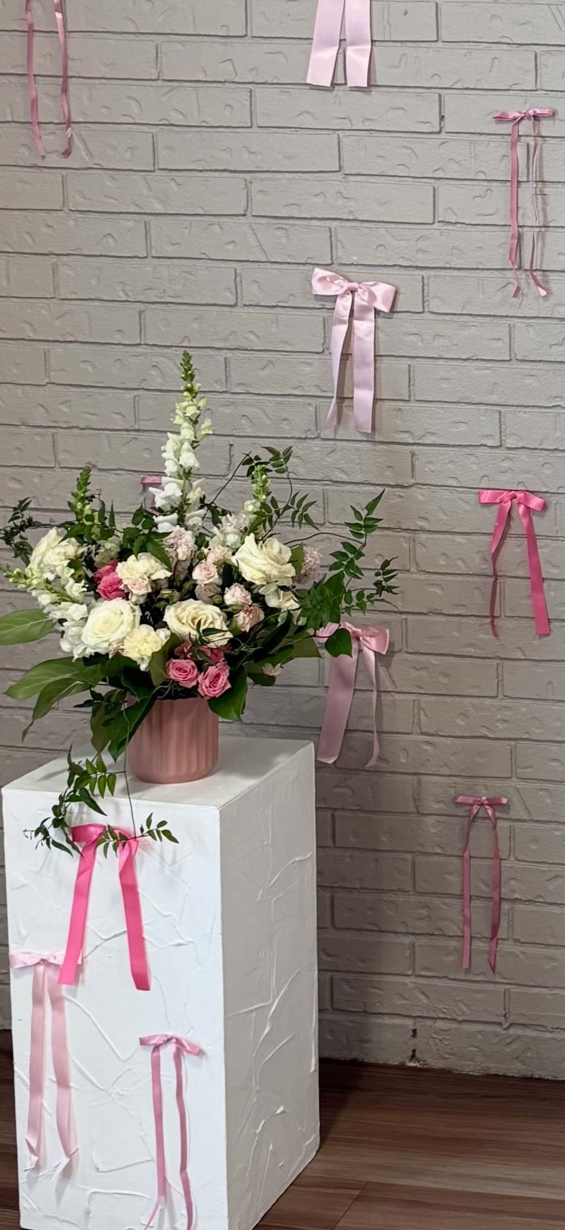 A floral arrangement with white and pink roses, white snapdragons, and greenery in a pink vase, placed on a white pedestal against a brick wall decorated with pink ribbons.