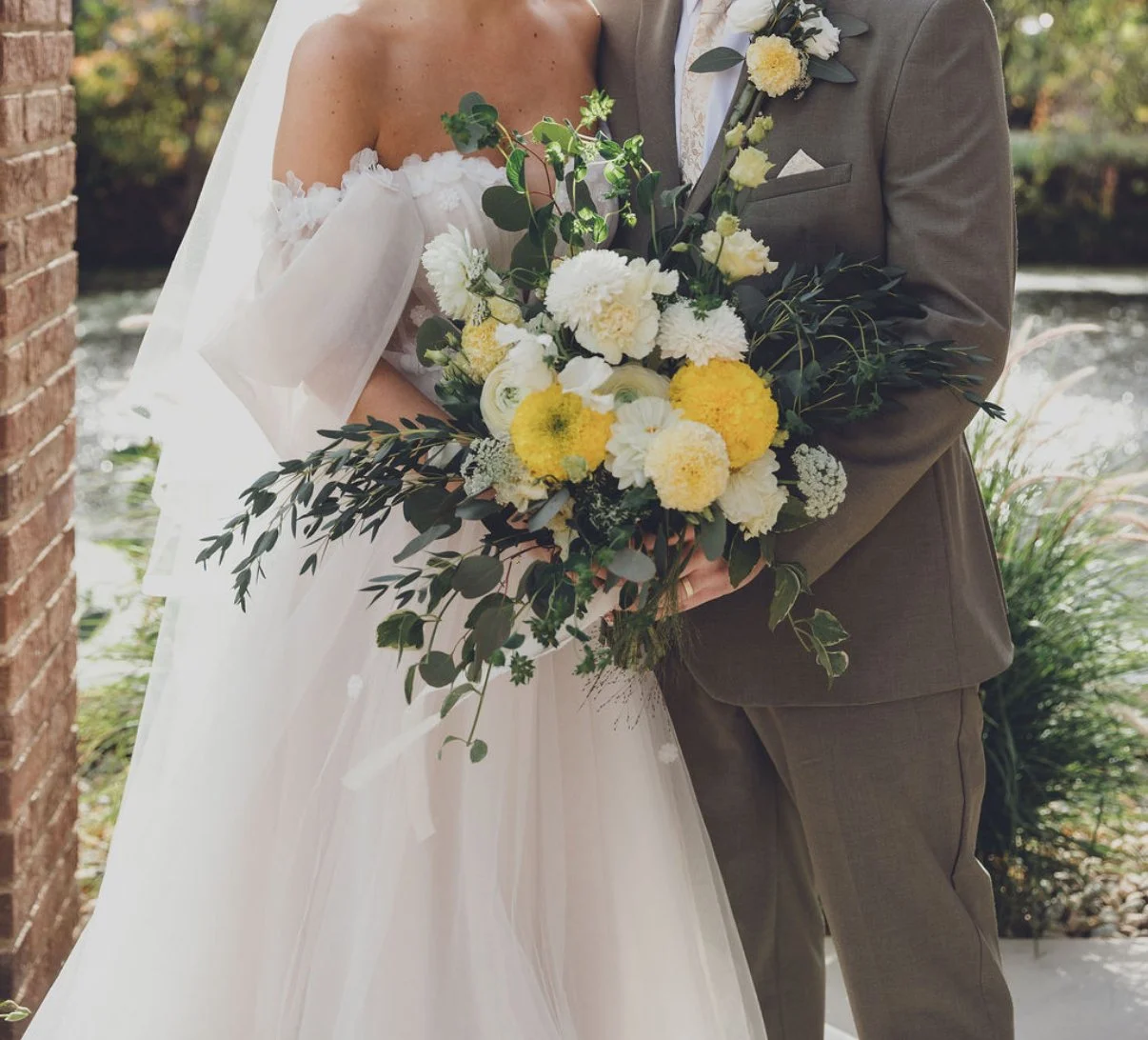 A bride and groom hold a large bouquet of white and yellow flowers with green foliage during their wedding.