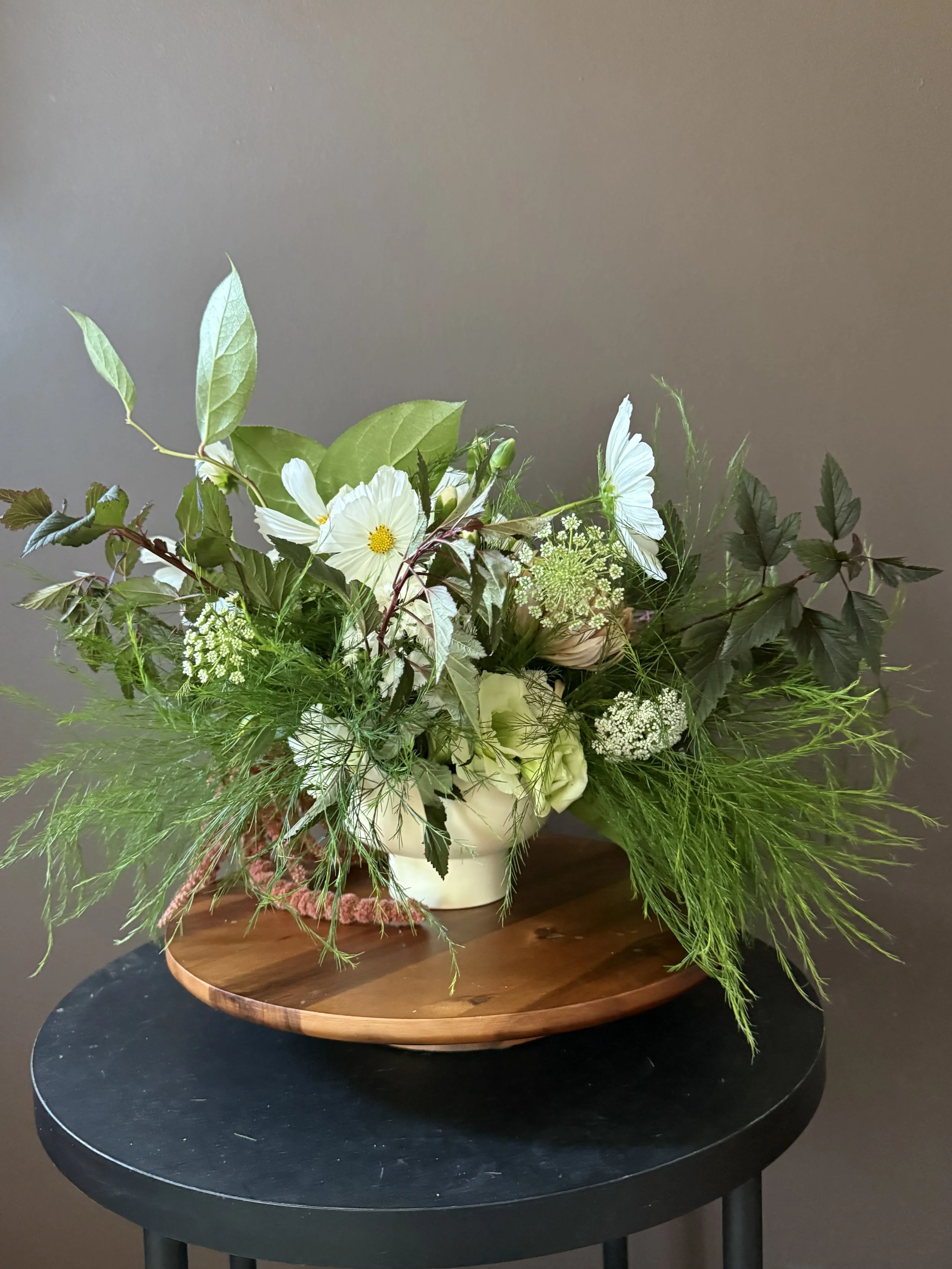 A floral arrangement in a white vase on a round wooden tray, featuring white flowers, green foliage, and delicate white herbs.