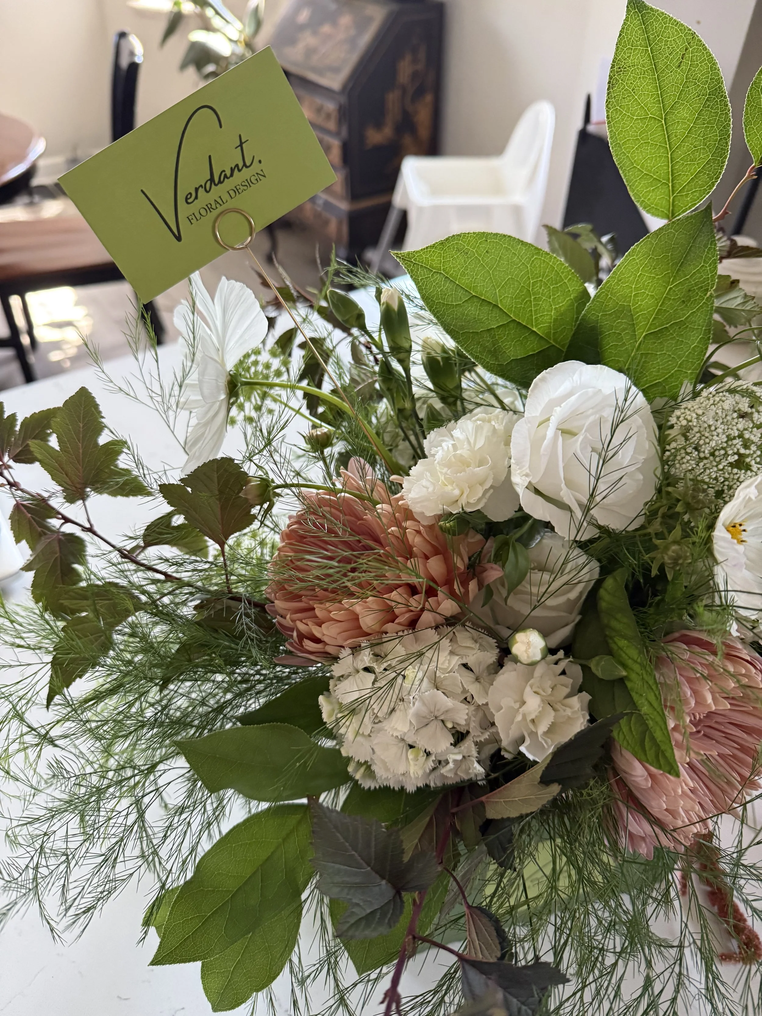 A bouquet of white and pink flowers with green leaves and foliage on a table in a room, with a business card reading 'Verdant. Floral Design' inserted into the arrangement.