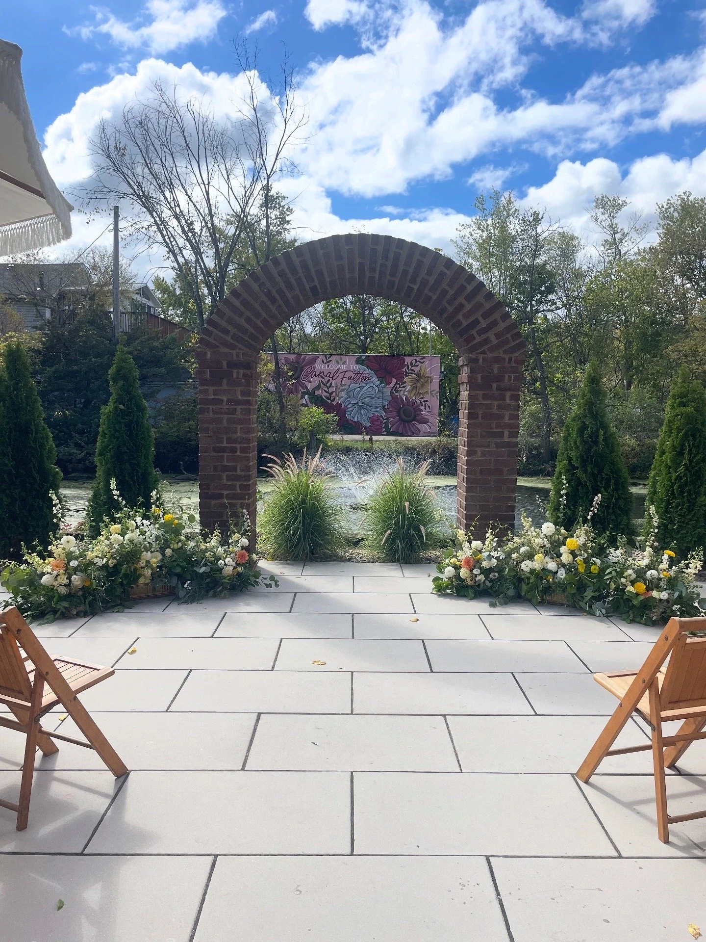 An outdoor patio area with white tile flooring, two wooden chairs, a brick archway, and decorative flowers along the sides. In the background, there is a pond with fountain, trees, and a colorful banner that says 'Welcome to Canal Fulton' under a partly cloudy sky.