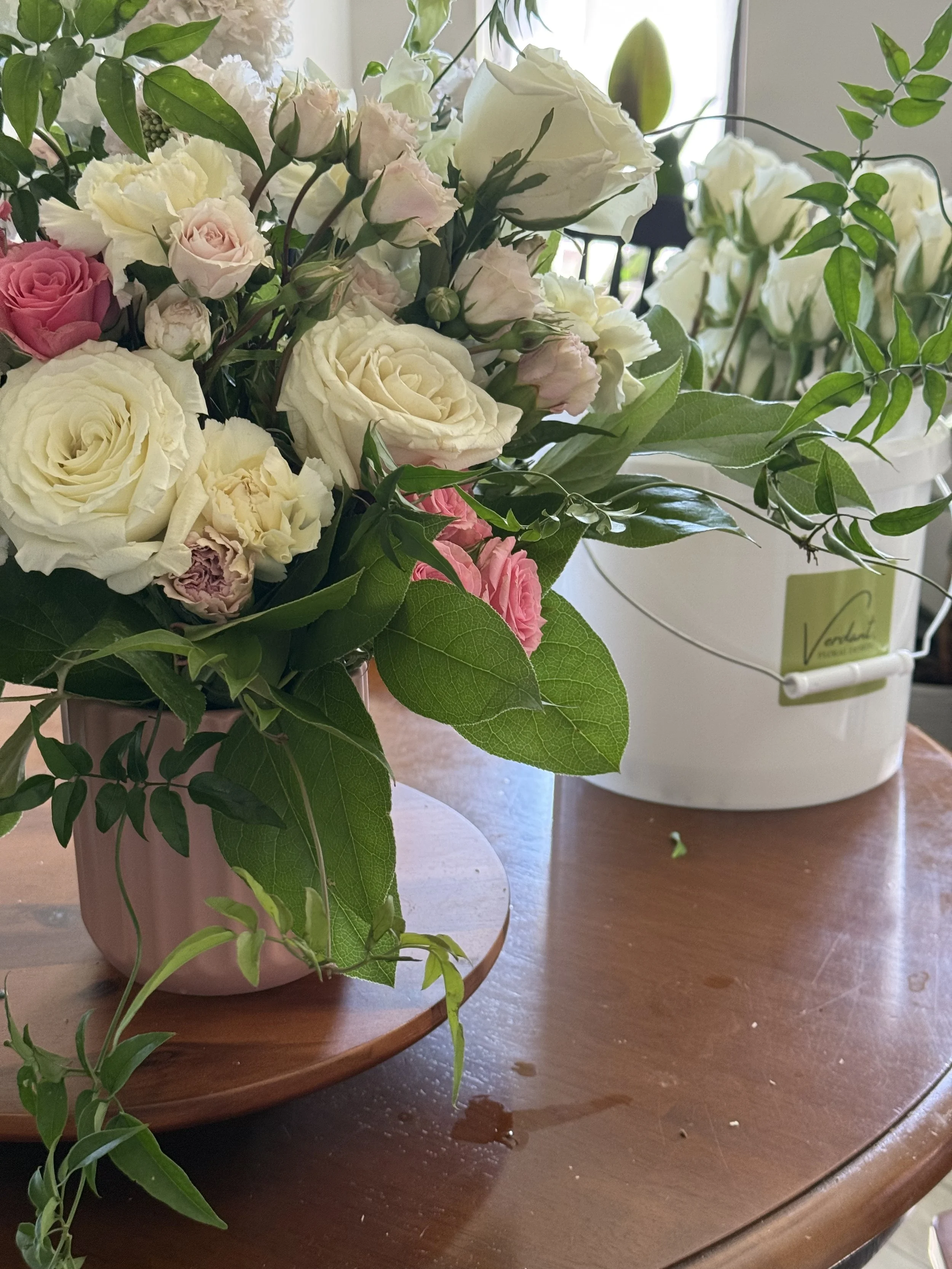 A floral arrangement with white, pink, and cream roses, and green foliage in a pink vase on a wooden table.