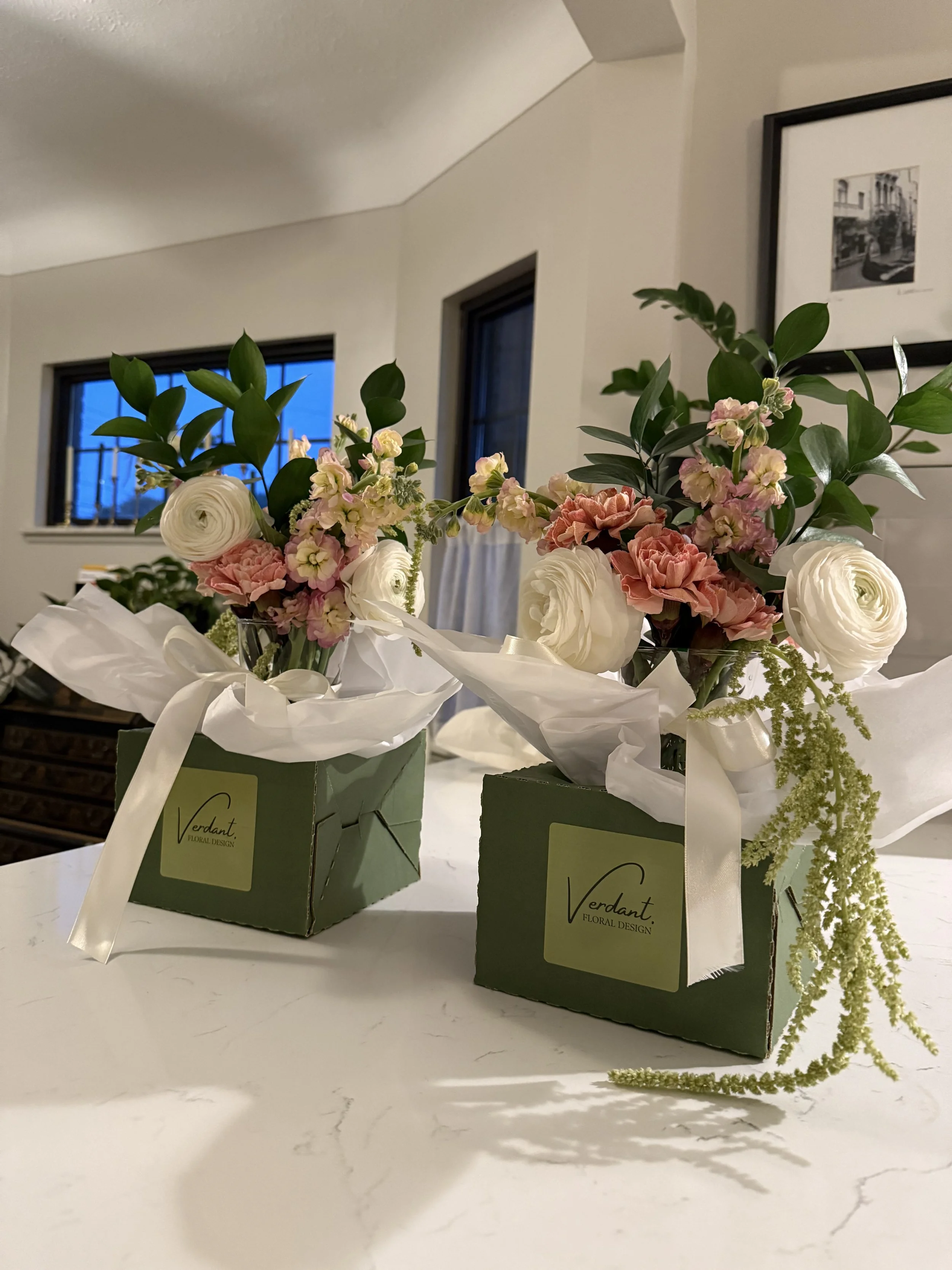 Two floral arrangements in green boxes with white tissue paper and white ribbons, featuring pink and white flowers with green leaves, placed on a white marble countertop in a modern home interior.