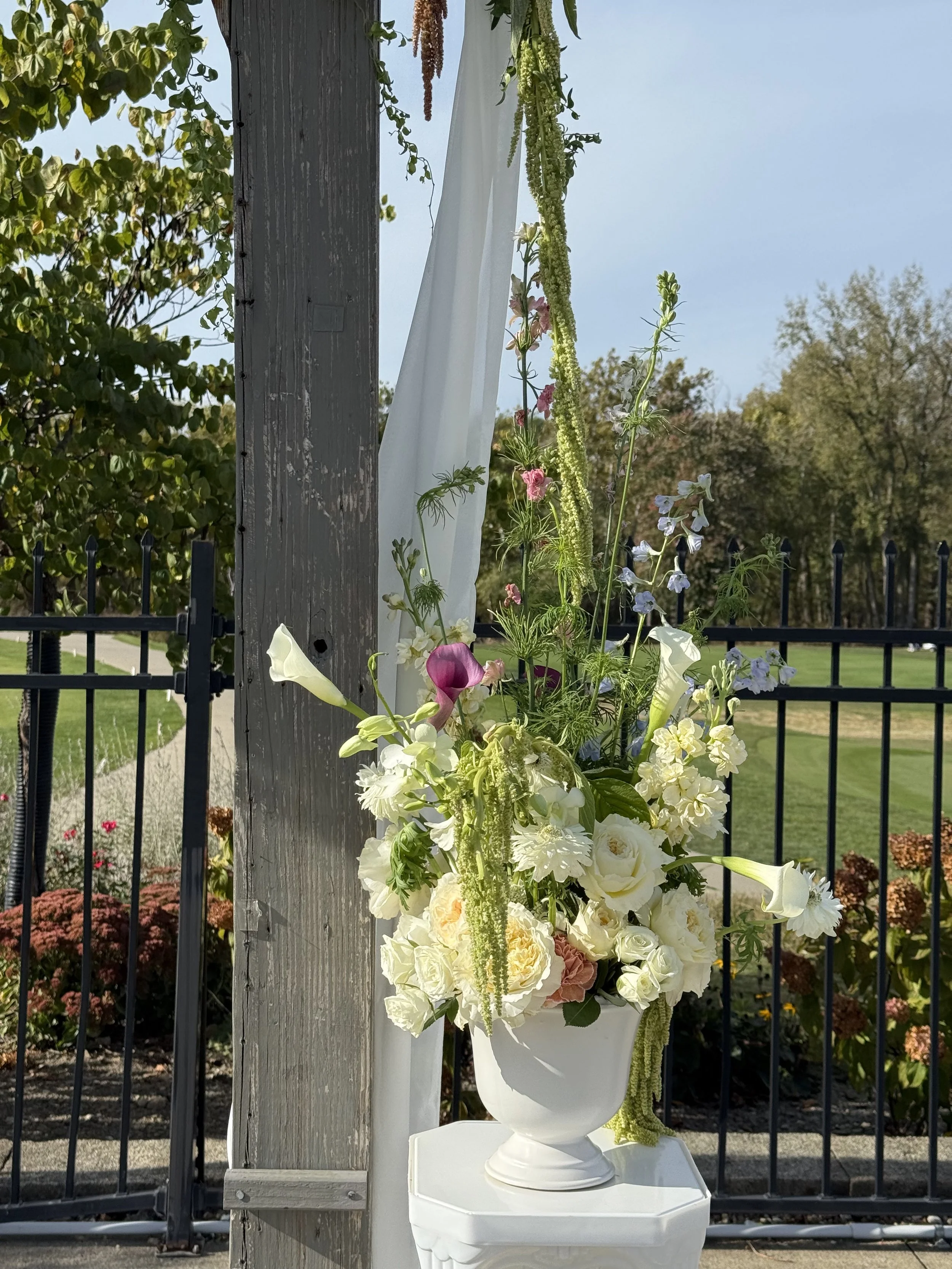 A white urn filled with a variety of flowers, including white roses, calla lilies, and other colorful blooms, set on a white pedestal outdoors with green trees and a black fence in the background.