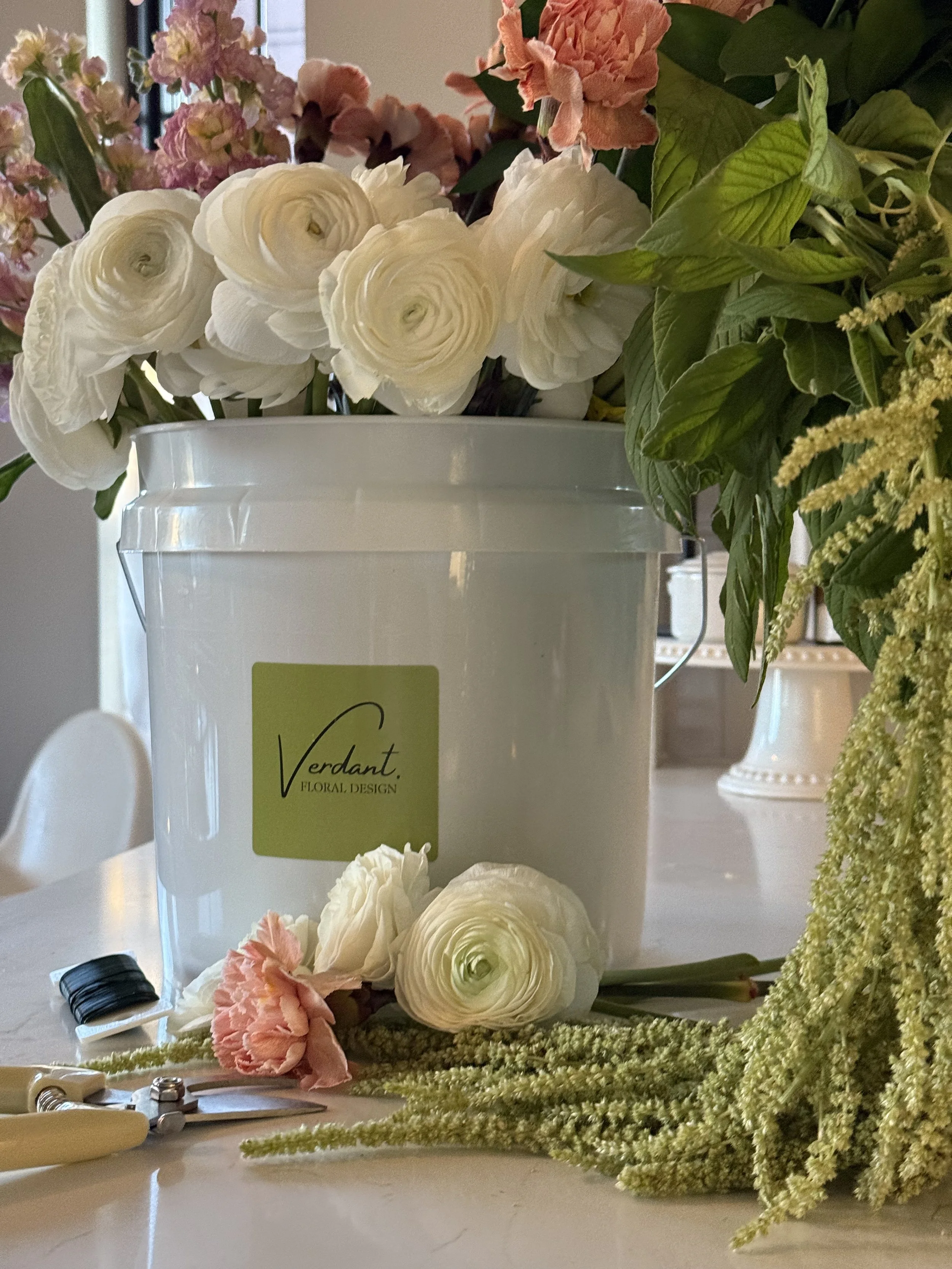 A white bucket with a green label reading 'Verdant Floral Design' holding white and light pink flowers, placed on a table with some flowers and tools nearby.