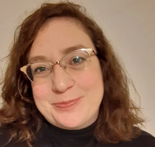 A woman with curly red hair and glasses smiling at the camera.