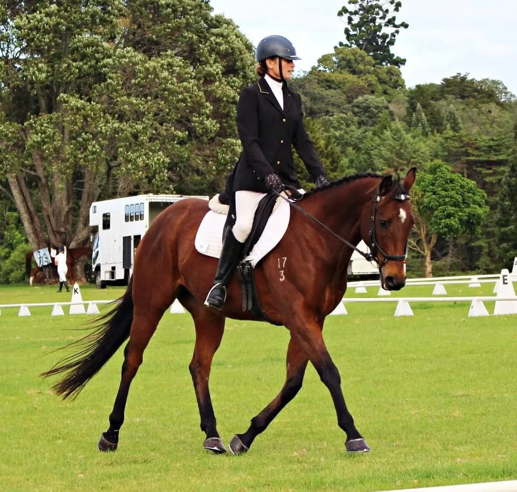 A woman dressed in formal riding attire, including a helmet and gloves, riding a brown horse on a grassy field during an equestrian event. There are white dressage arena markers and another horse and rider in the background.
