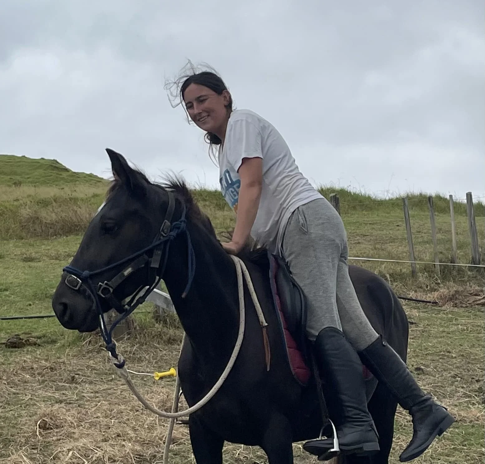 Woman riding a black horse outdoors on a cloudy day, wearing gray pants and black riding boots, with wind blowing her dark hair.