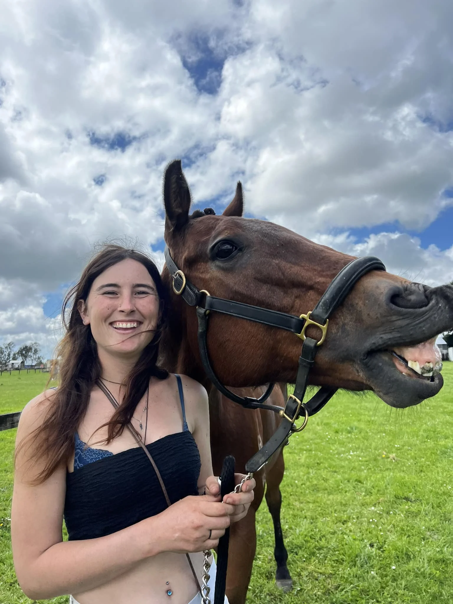 A smiling woman in a black top standing next to a brown horse outdoors on a grassy field with cloudy sky in the background.