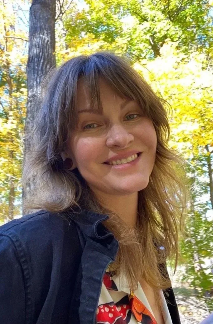 A woman with wavy, light brown hair stands outdoors in a forested area with green and yellow autumn leaves. She is smiling and wearing a black jacket over a patterned shirt.