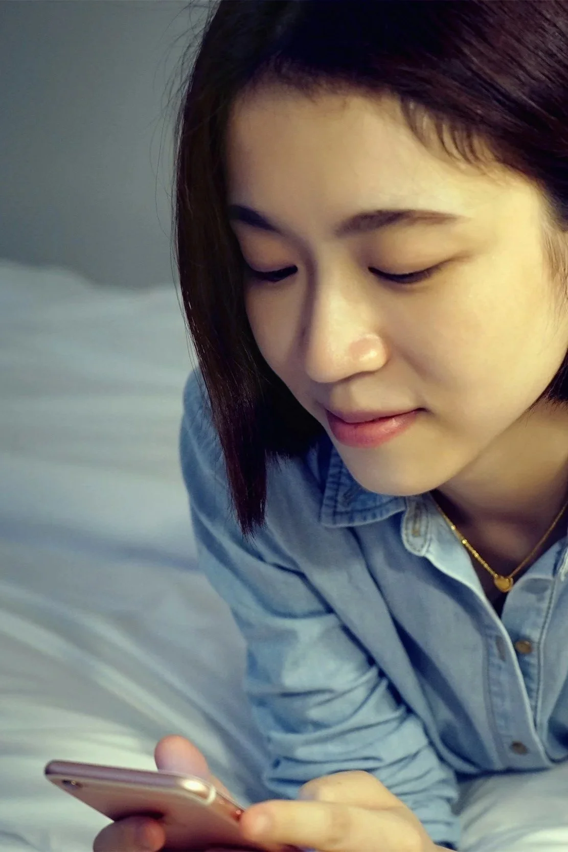 Young woman with shoulder-length hair, wearing a denim jacket and a gold necklace, looking at her smartphone.