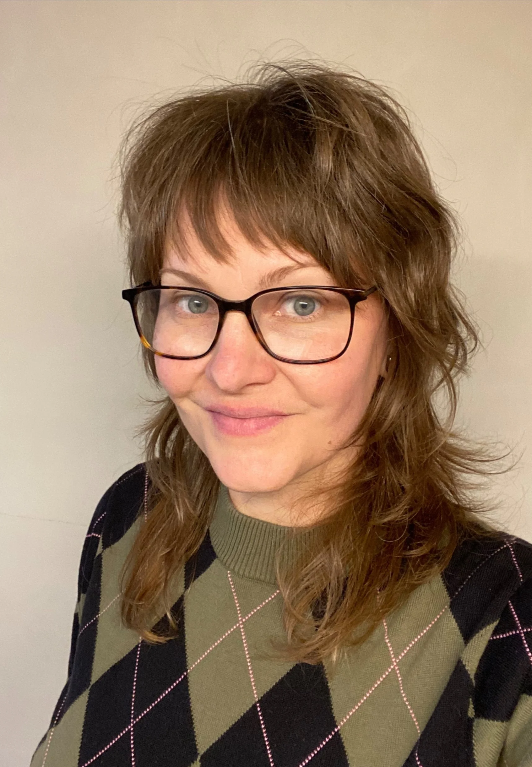 A woman with light skin, shoulder-length wavy brown hair, glasses, and a subtle smile, wearing a green and black argyle sweater with pink stitching.