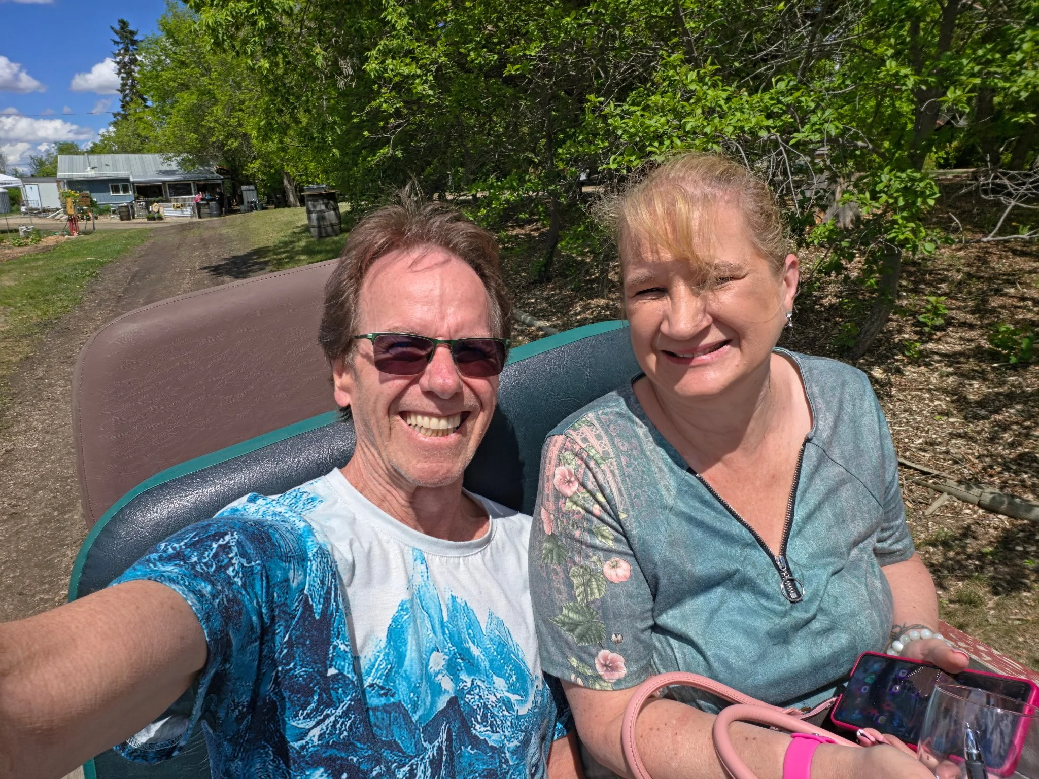 Two women taking a selfie outdoors with green trees and a blue sky in the background. One woman wears glasses and a colorful blue shirt, the other has blonde hair and wears a patterned top. There are some personal items like a phone, purse, and drinks on a table nearby.
