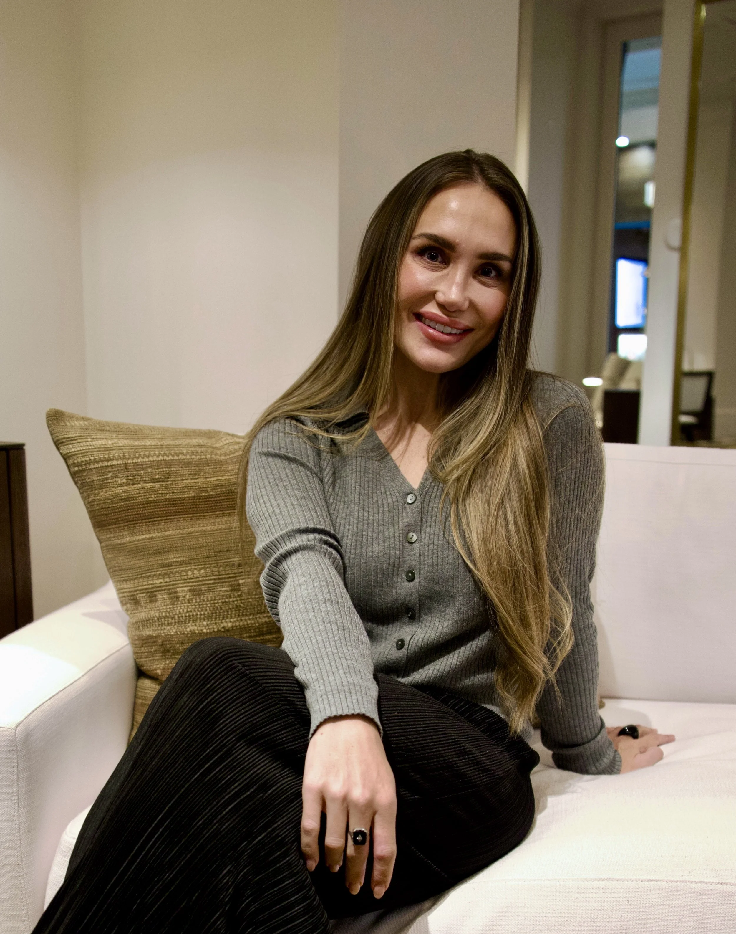 A woman with long brown hair sitting on a white couch in a living room, smiling at the camera, wearing a gray ribbed long-sleeve top and black pants.