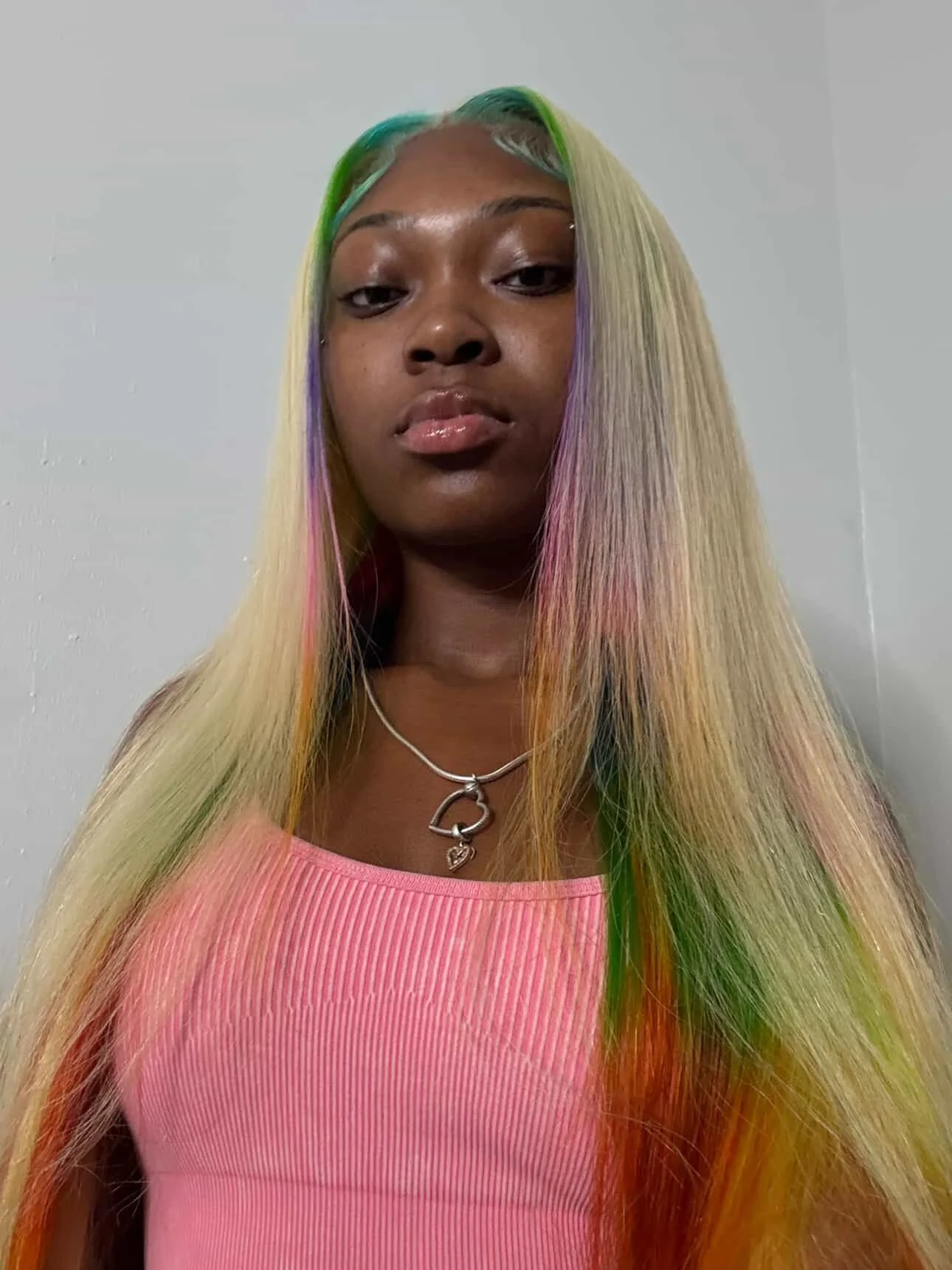 Young woman with long, multicolored hair in shades of blonde, pink, green, and purple, wearing a pink ribbed tank top and a silver necklace with a heart pendant.