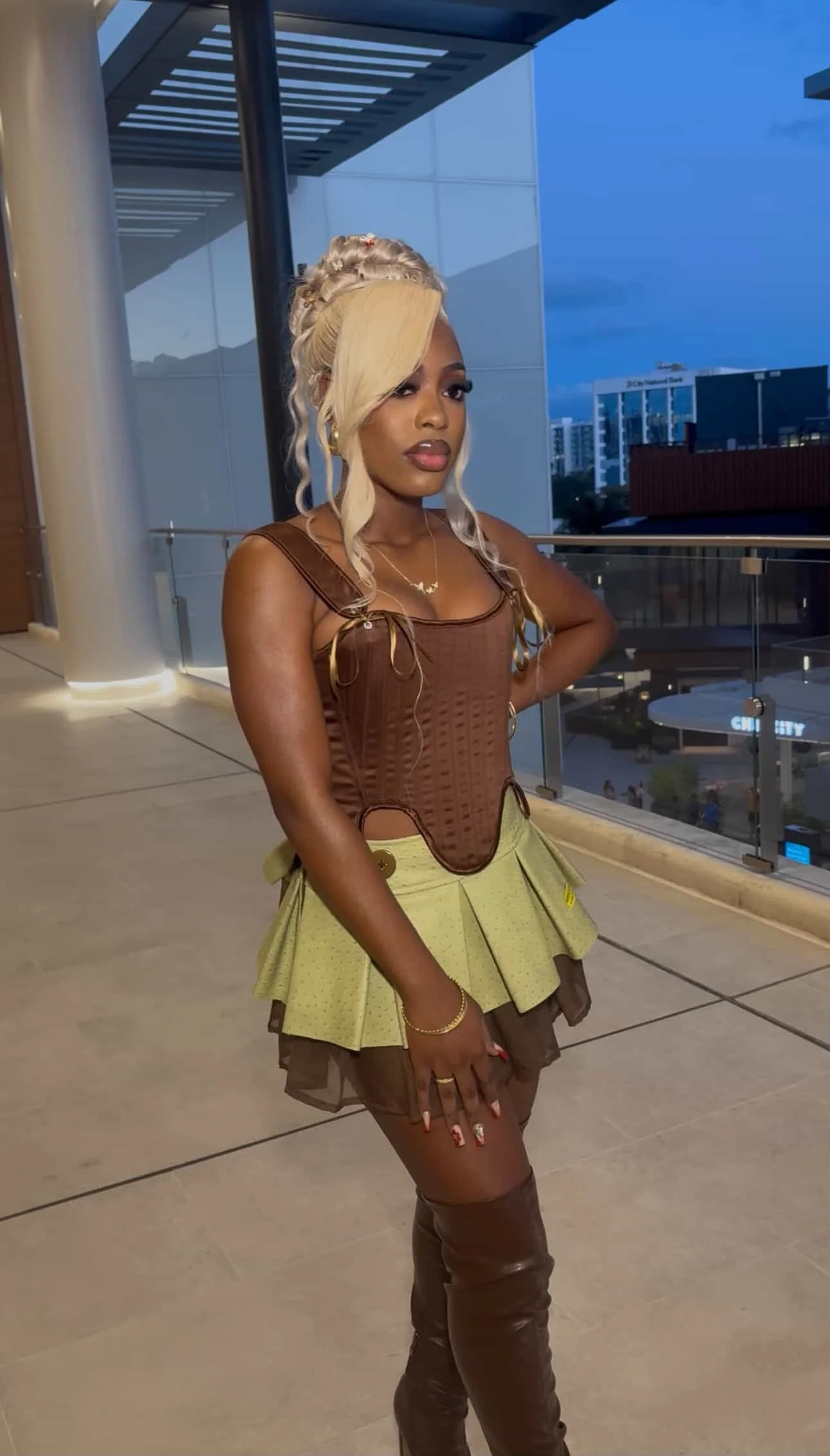 A woman with blonde hair styled in loose waves and a side-swept bang, wearing a brown corset top, a light yellow skirt with pleats, brown thigh-high boots, and jewelry, standing on a balcony with city buildings in the background during dusk.