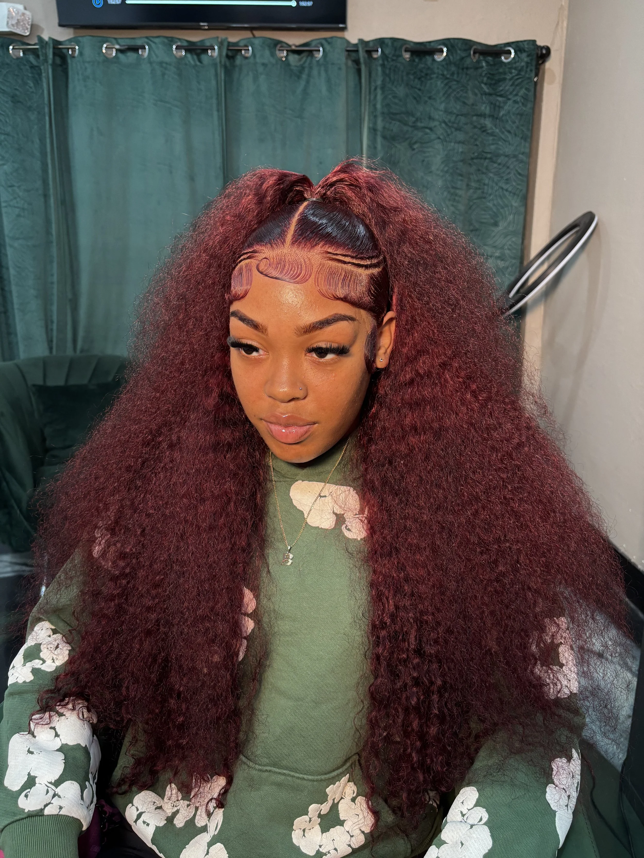 Young woman with voluminous, curly burgundy hair styled in a half-up, half-down hairstyle, with intricate baby hair designs and sleek edges, wearing a green floral-patterned top and sitting indoors.