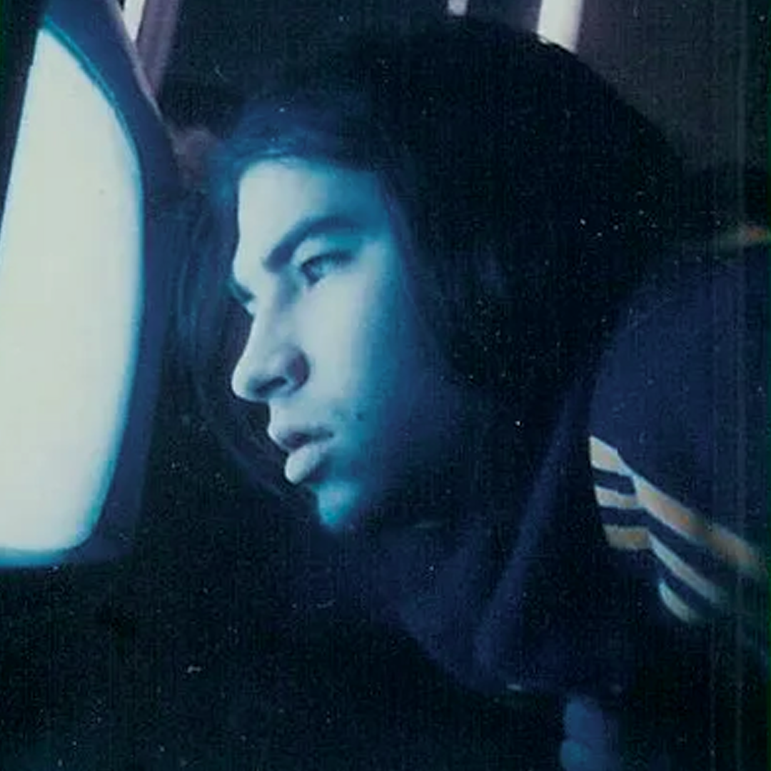 Close-up of Canadian indie artist Spookey Ruben with dark hair, gazing into an analog TV screen, wearing a dark jacket with white striped sleeves.