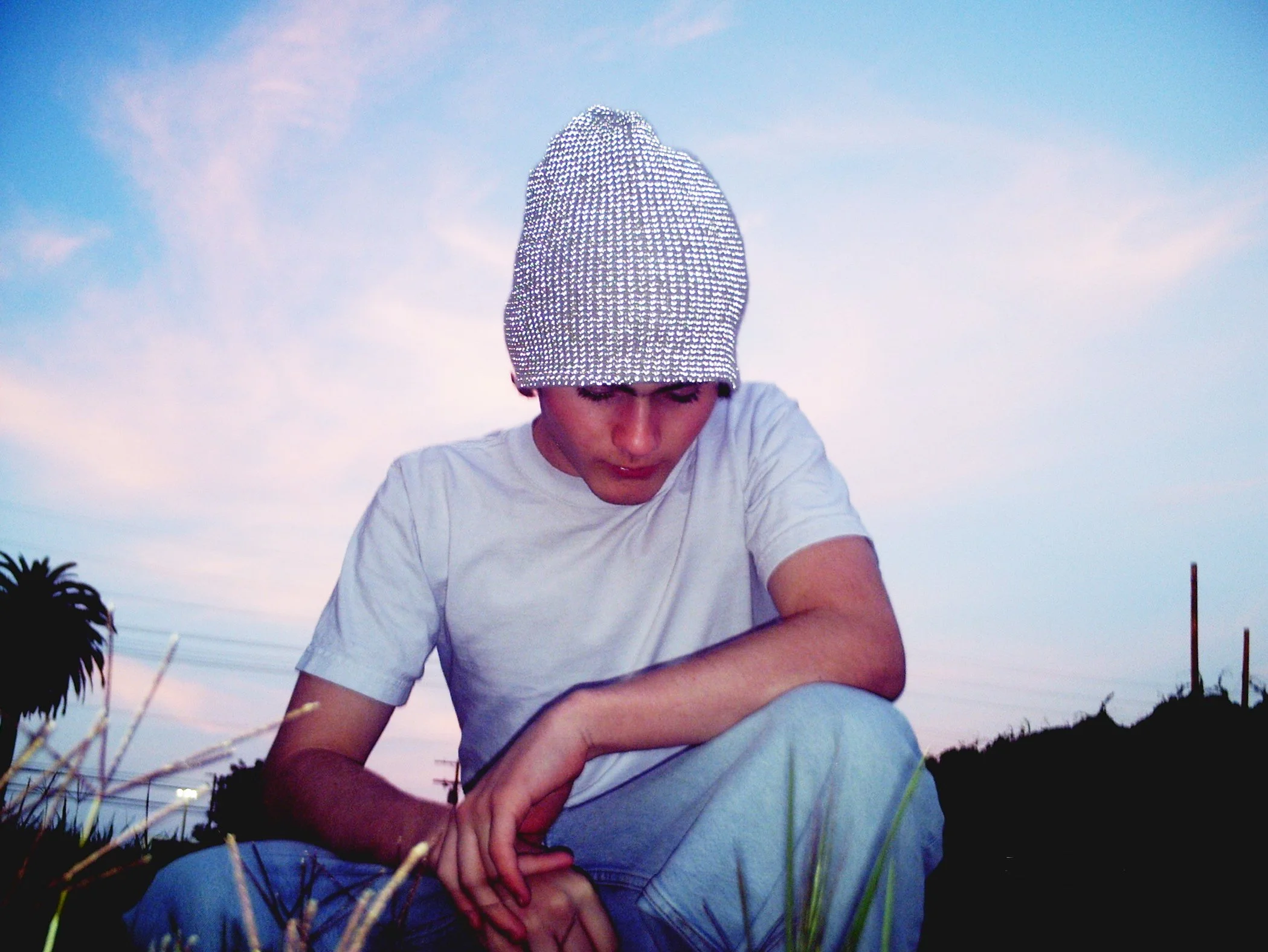 LA-based artist Gus Blu wearing a hat with a cloudy sky in the background, looking downward, sitting outdoors.