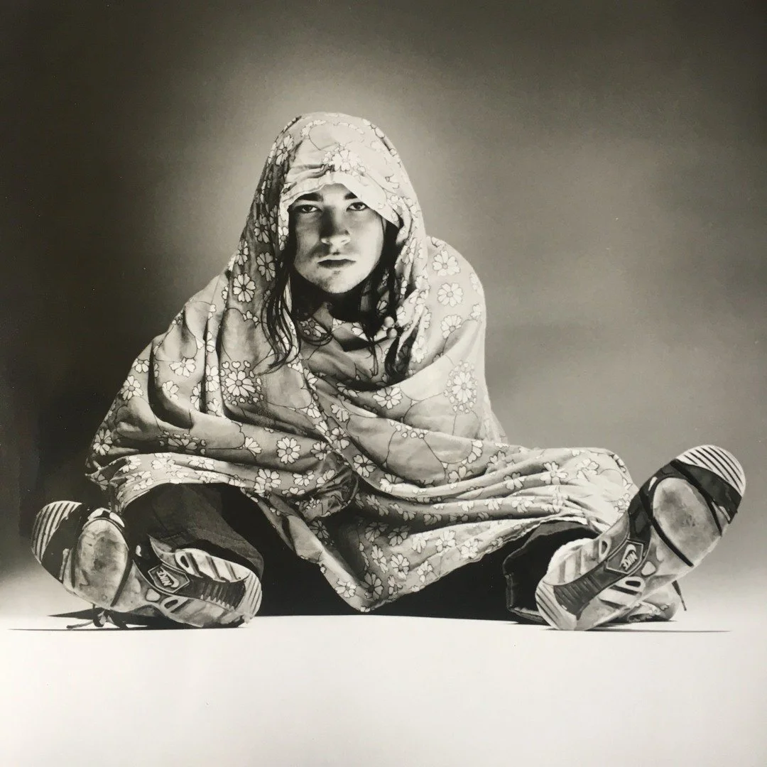 Black-and-white photo of Canadian indie artist Spookey Ruben sitting on the floor, wrapped in a floral-patterned blanket, wearing sneakers, with a serious expression.