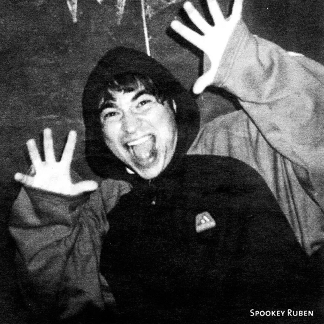 Canadian indie artist Spookey Ruben smiling with his mouth open, wearing a hat and a jacket, with both hands raised in a playful gesture.