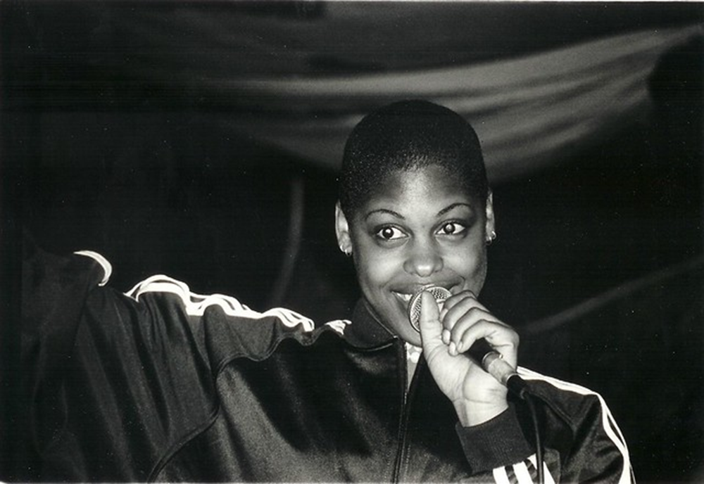Black and white photograph of American hip-hop artist T-Love with short hair, holding a microphone and smiling, wearing a sport jacket with white stripes on sleeves.