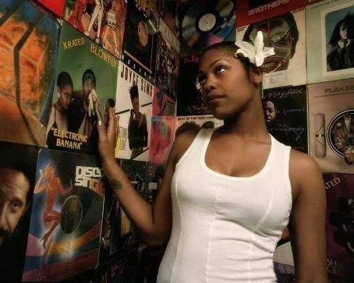 American hip-hop artist T-Love with a white flower in her hair, wearing a white tank top, standing in front of a wall covered with vinyl records and album covers, placing her hand on one of them.