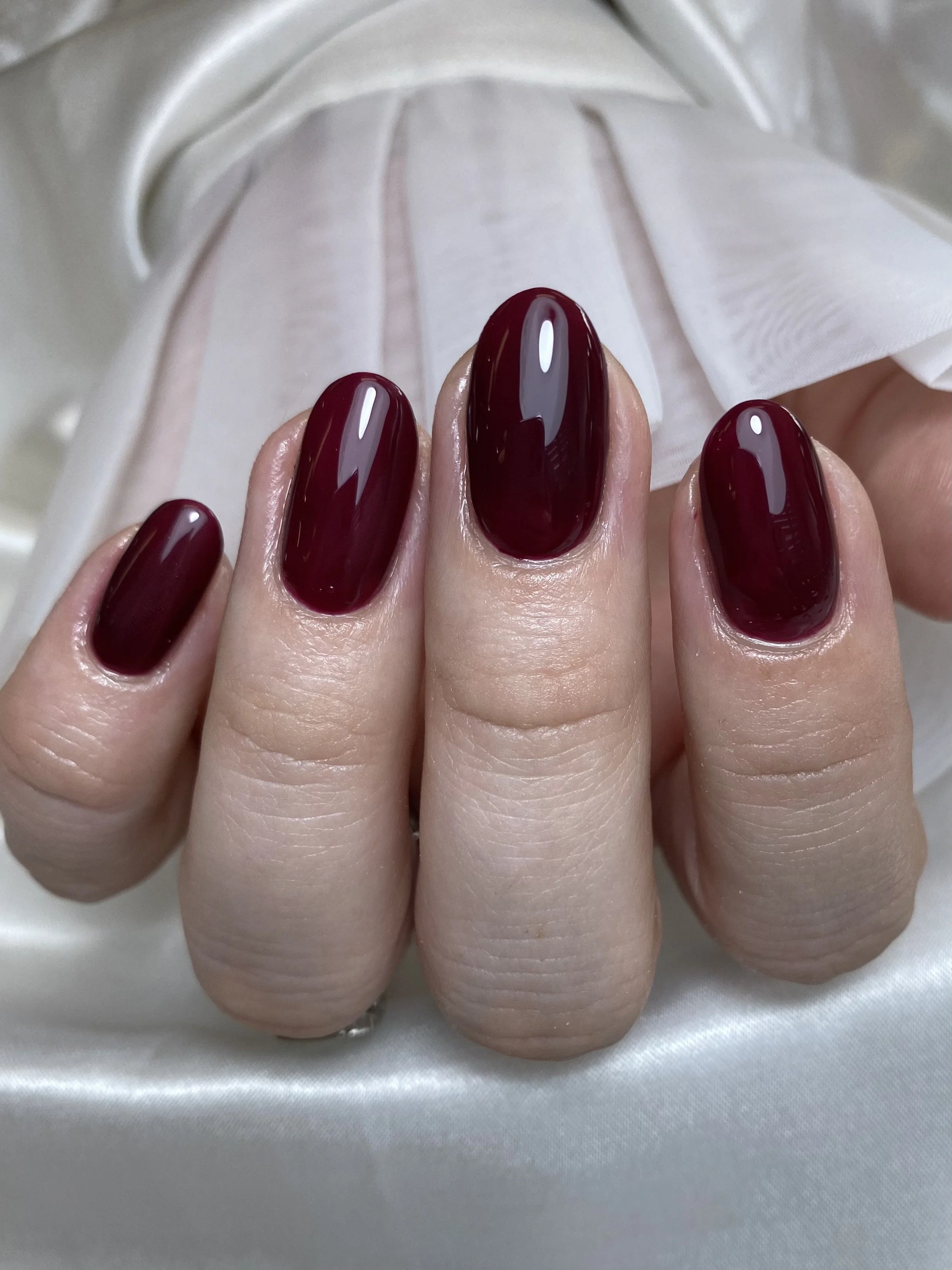 Close-up of a hand with manicured nails painted in dark red, set against a silky, light-colored fabric background.