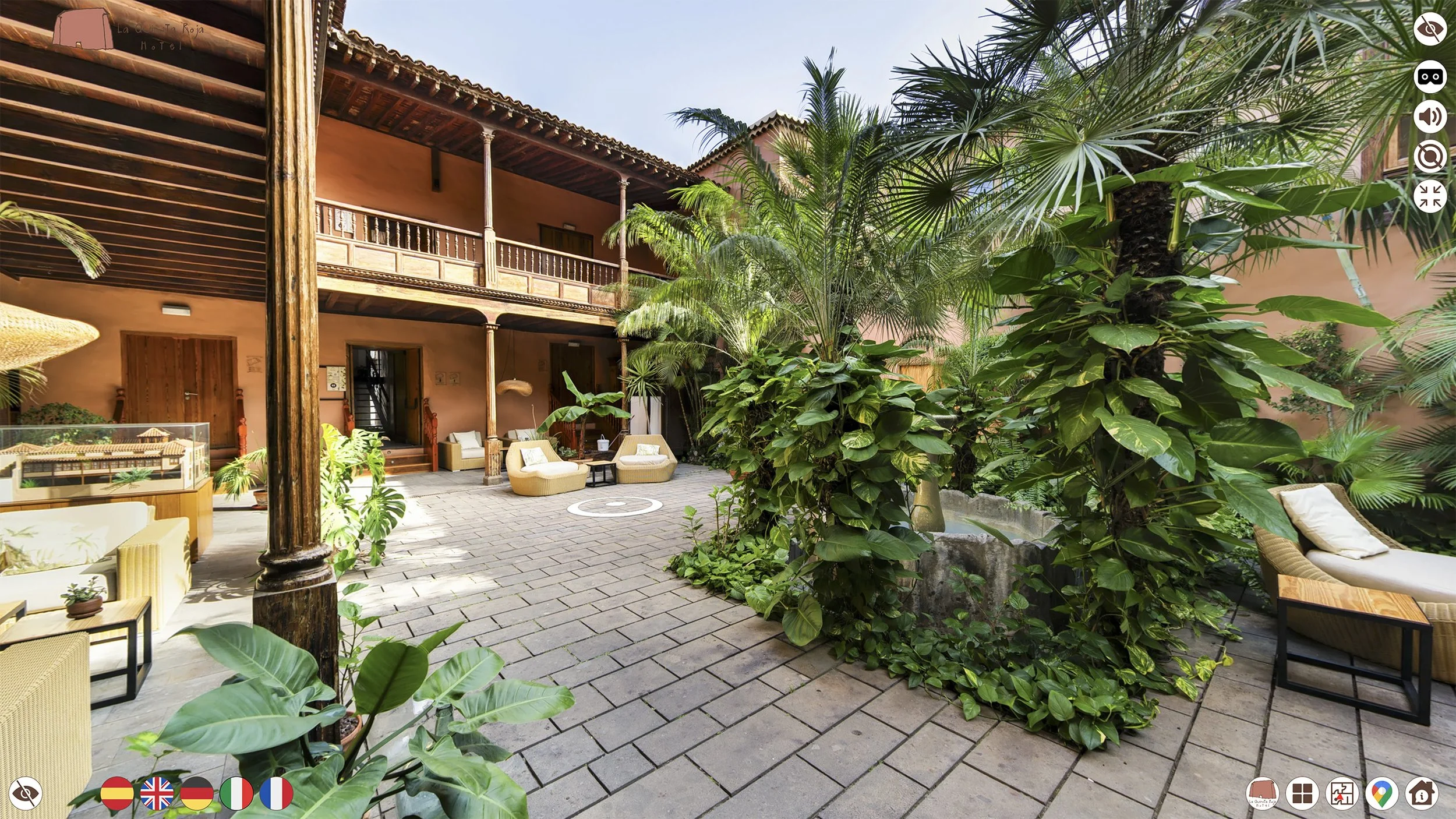 A spacious hotel courtyard with tropical plants, patio furniture, and a two-story building with a balcony. The area is paved with gray stones and decorated with outdoor seating.
