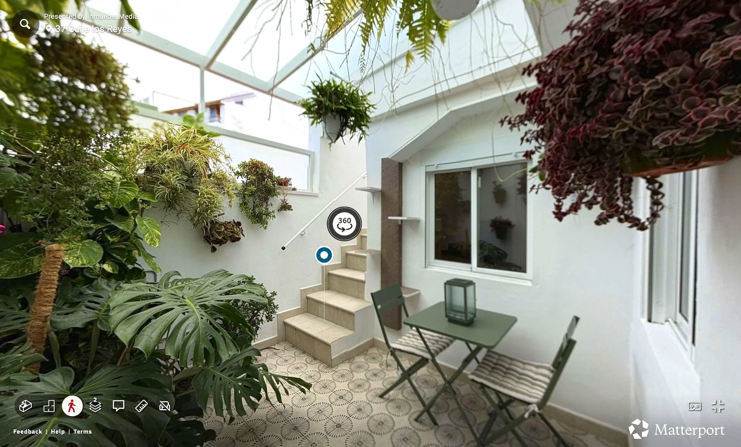 Jardín cubierto con varias plantas en macetas colgadas en las paredes y en el suelo, una mesa con sillas y una linterna en la mesa, ventanas y escaleras en un espacio cerrado con techo de cristal.