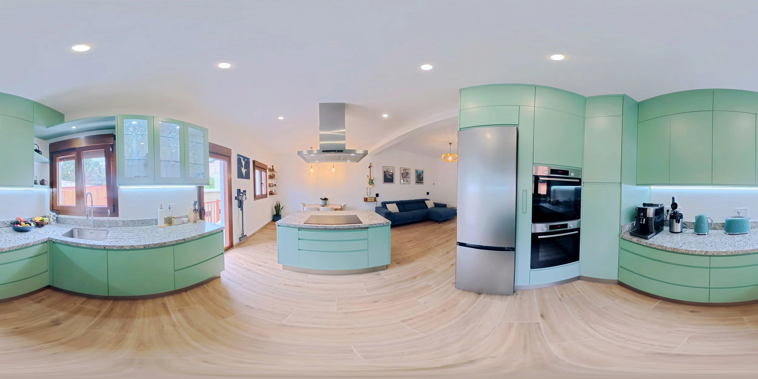 Panoramic view of a modern kitchen and living room with green cabinets, granite countertops, wooden flooring, and a blue sofa.