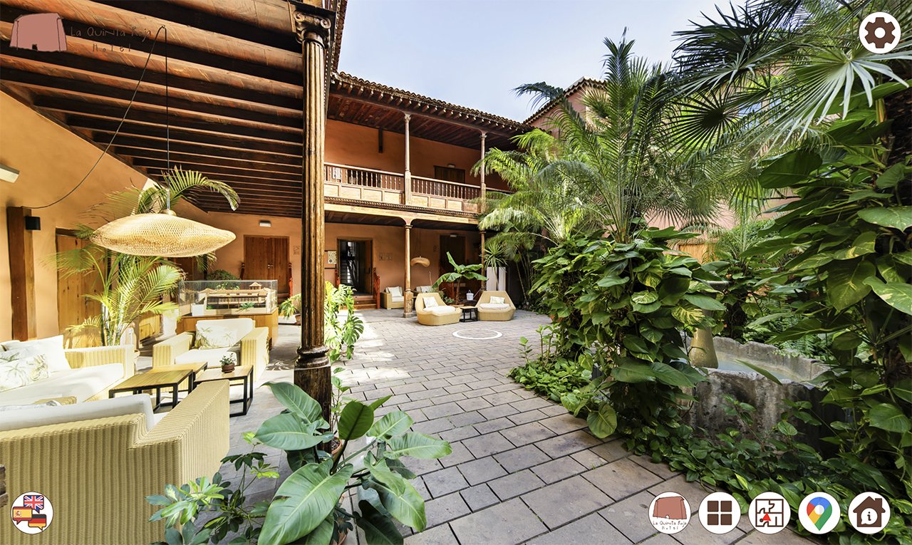A lush courtyard with tropical plants and outdoor furniture, featuring a two-story building with a wooden balcony and a tiled roof.