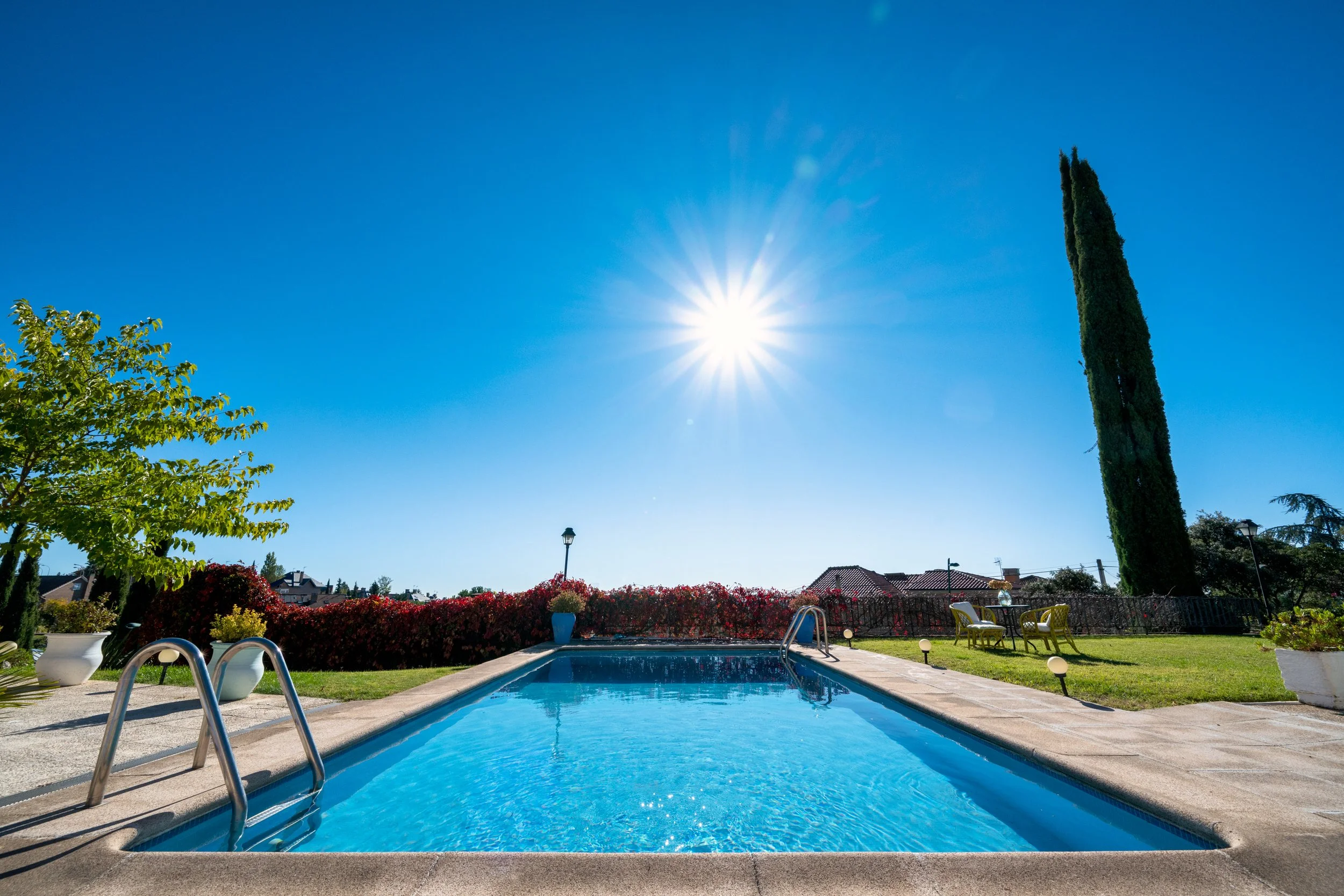 Piscina en un jardín soleado con árboles y sillas, cielo azul y sol brillante.