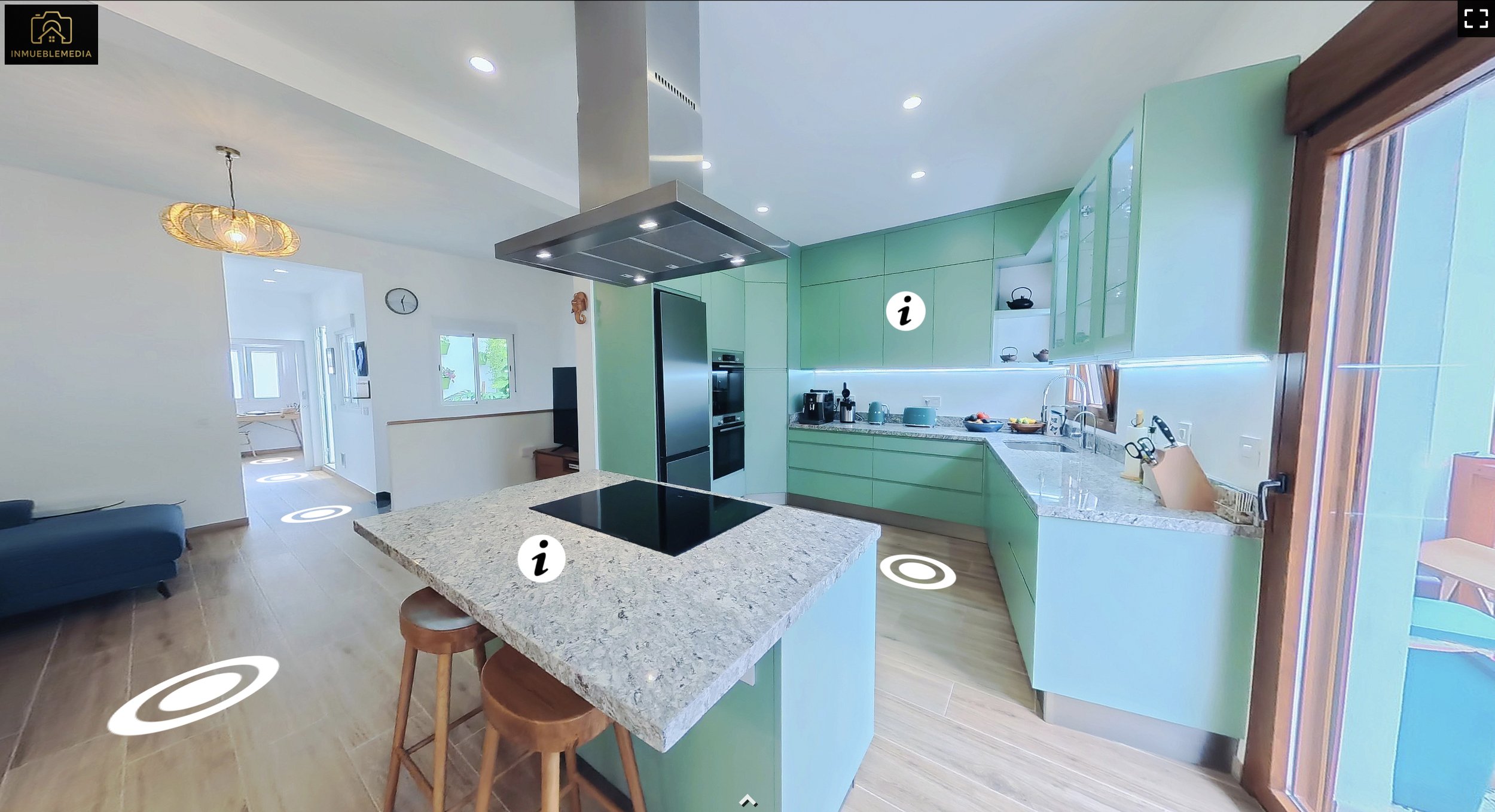 Cocina moderna con gabinetes de color verde, encimera de granito, estufa eléctrica en la isla, y ventana que da a un área exterior. Área de comedor y sala en el fondo.