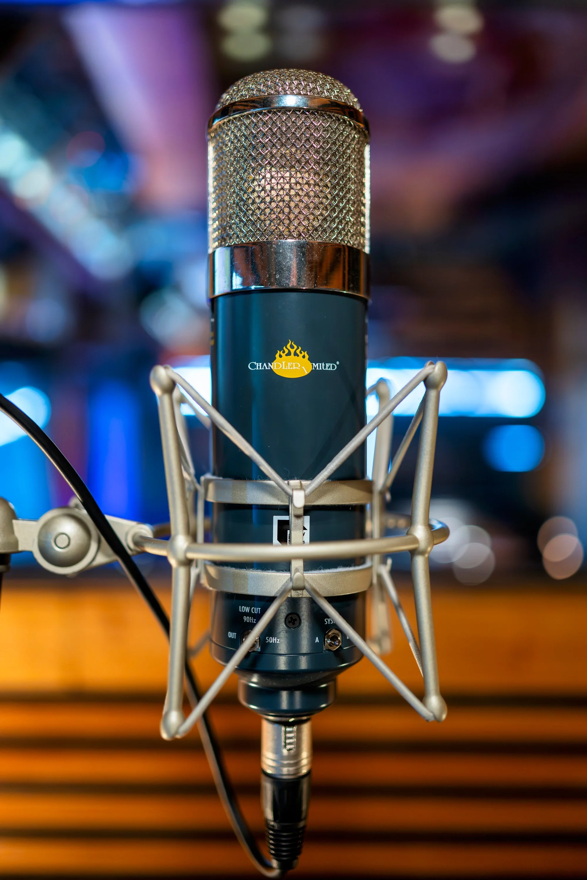 Un micrófono de estudio en primer plano con soporte y cables, fondo difuminado con luces de colores.