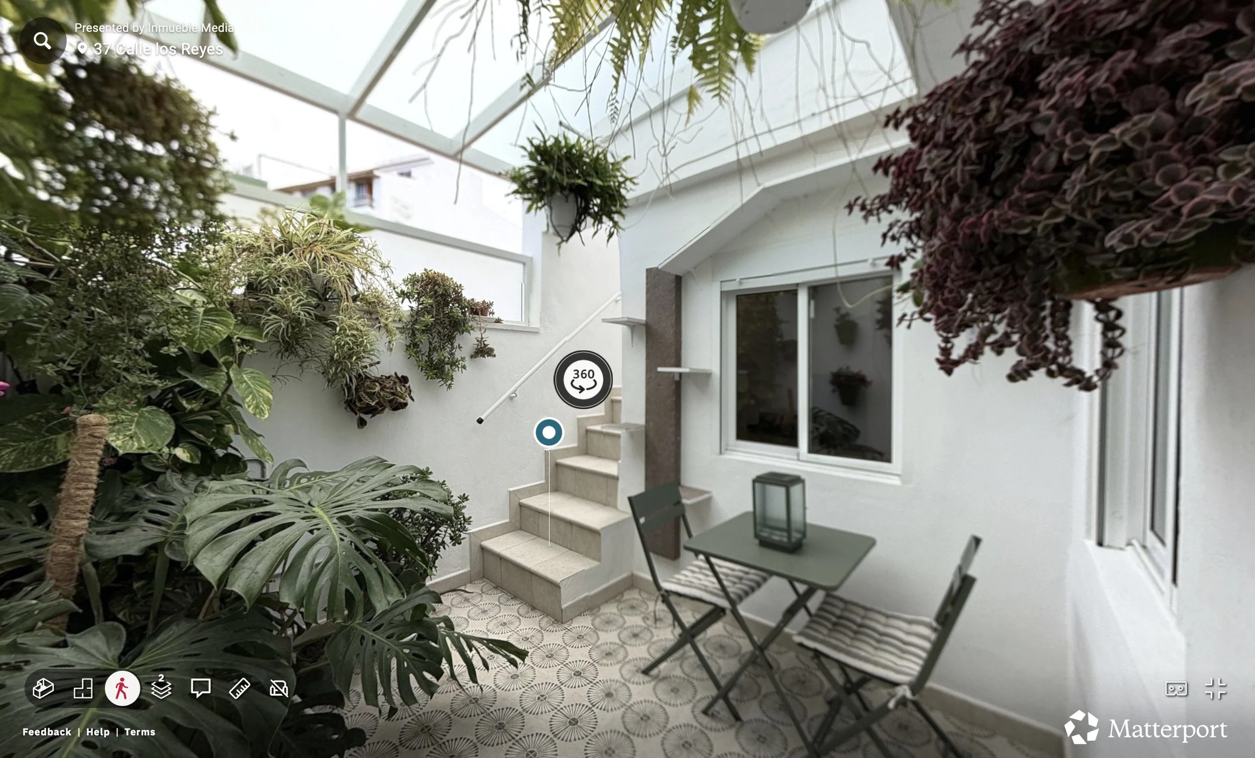 Patio con plantas verdes, mesa con dos sillas, ventanas blancas, y escaleras en una casa con techo de vidrio.